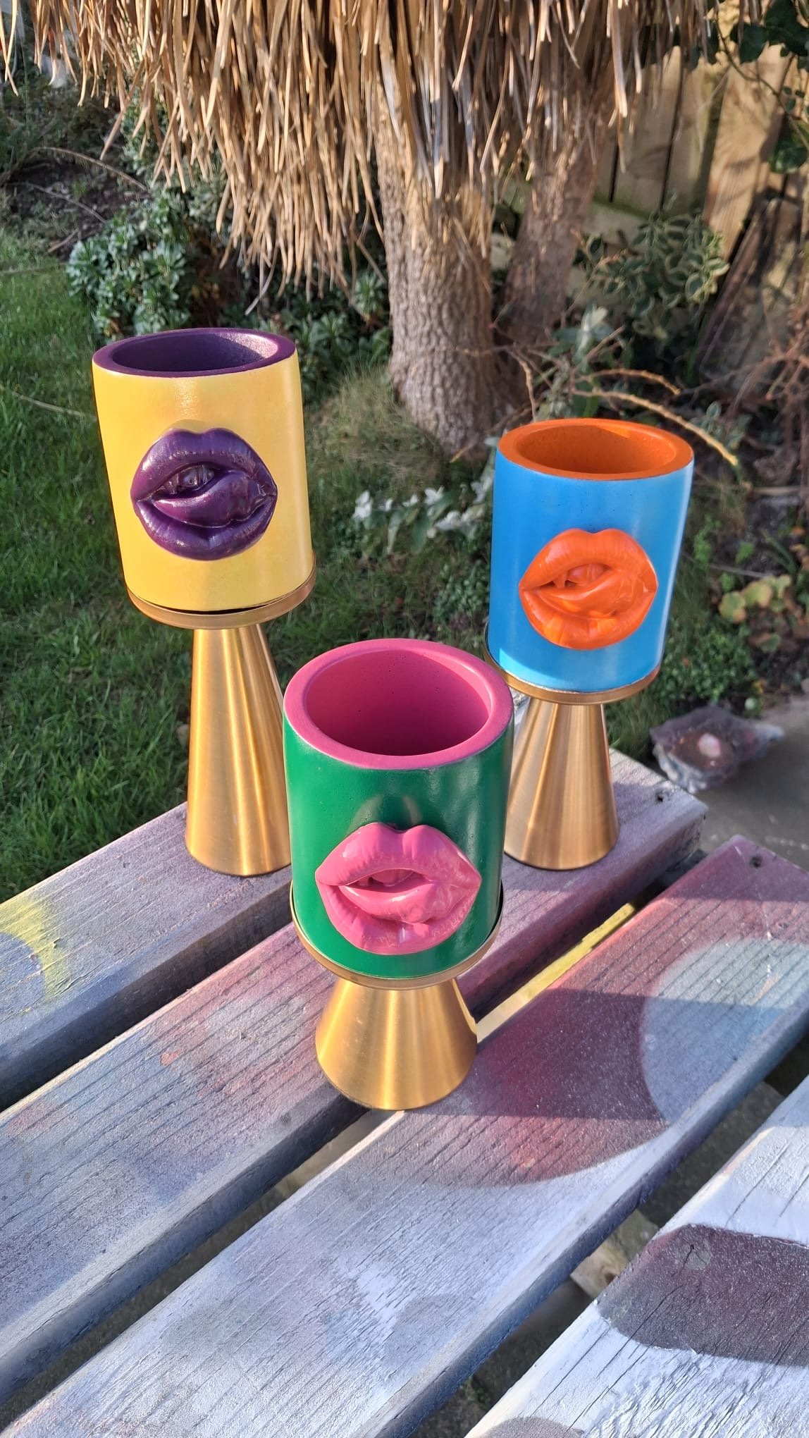 Three colorful decorative cups with lips and tongue designs on them placed on a wooden bench outdoors, with a grass and garden background.