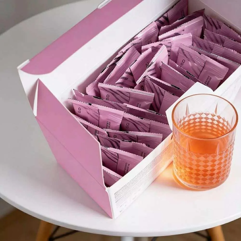 A pink box filled with individually wrapped pink packets next to a textured glass of orange-colored beverage on a white round table.