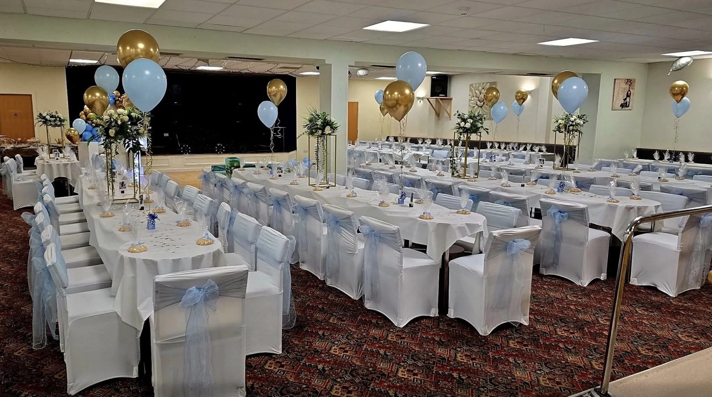 A banquet hall decorated for an event with tables covered in white tablecloths, white chairs with white covers and light blue bows, and balloon centerpieces in gold and light blue. There are floral arrangements and a stage in the background.