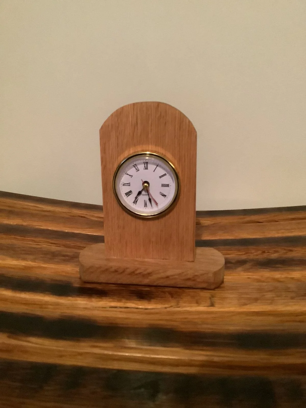A small wooden clock with a round white face, Roman numerals, and black hands, sitting on a wooden surface against a plain wall.