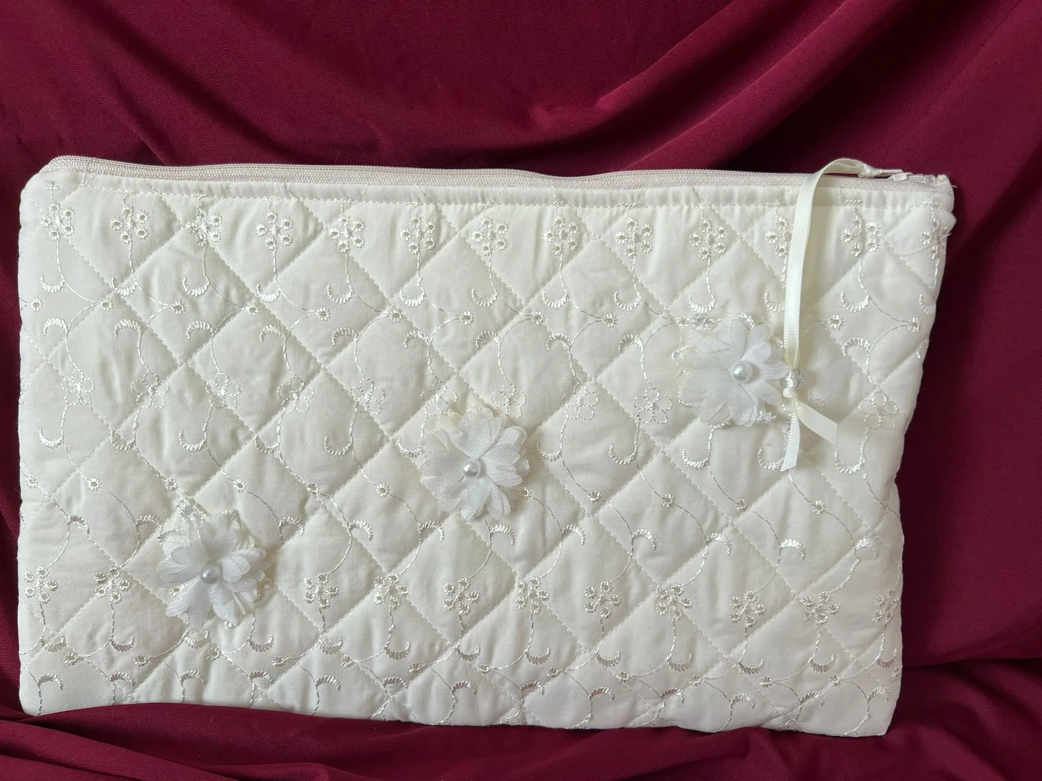 A white quilted fabric bag with embroidered floral patterns, decorated with three fabric flowers and pearl centers, and a satin ribbon bow, on a maroon fabric background.
