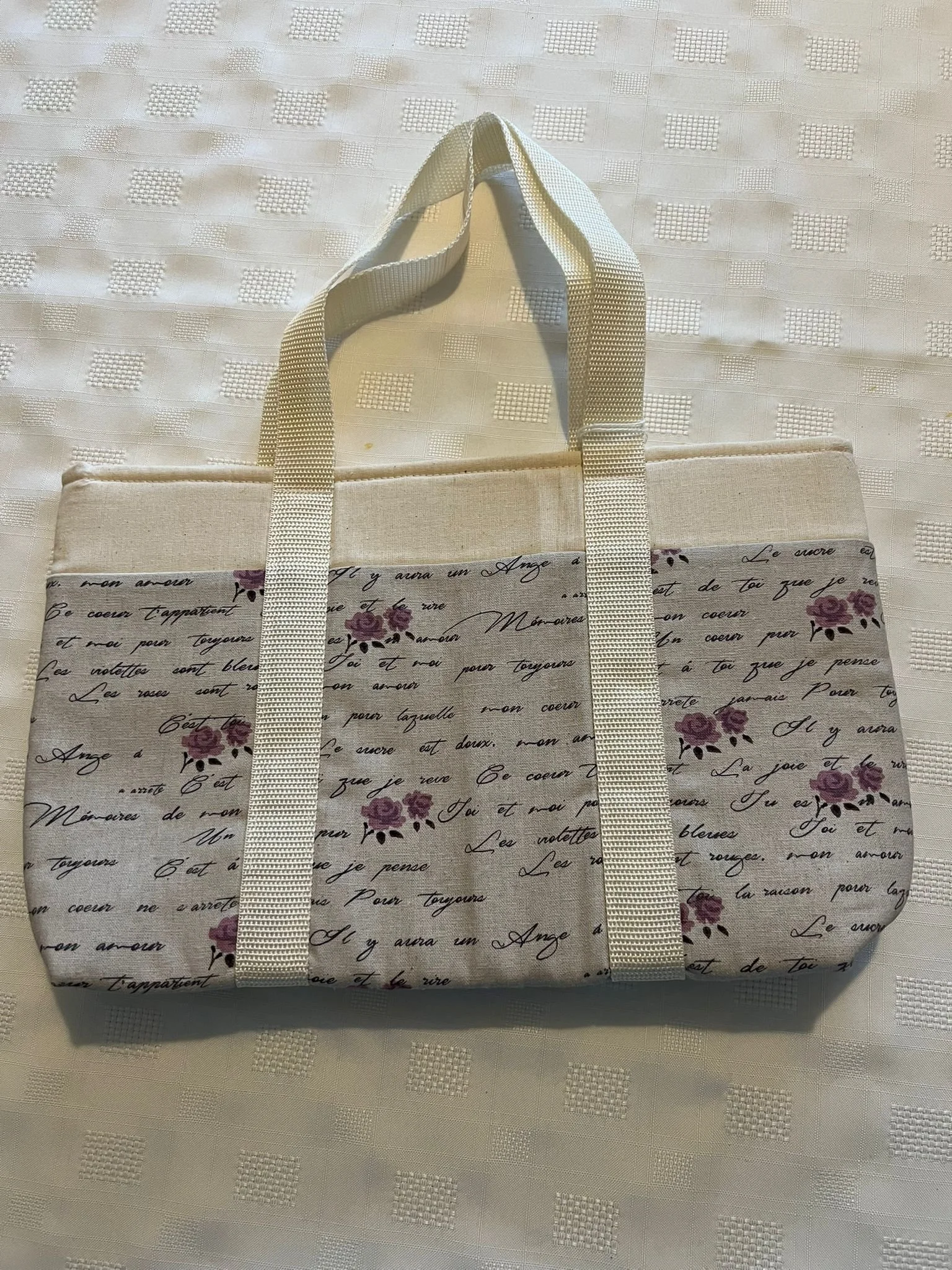 A fabric tote bag featuring French script and purple roses on a white background, with white straps, placed on a white textured tablecloth.