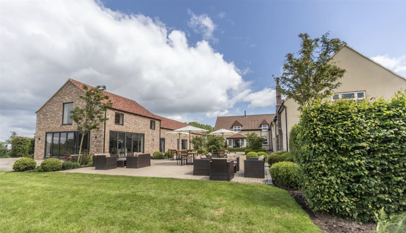 A well-maintained backyard with green grass, outdoor seating with umbrellas, and surrounding houses under a partly cloudy sky.