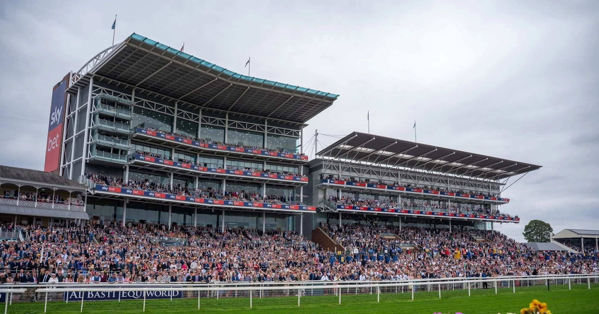 A large crowd of spectators at a horse racing event in front of a multi-story grandstand with advertisements for Sky Bet and a large red and blue banner for Sky Bet, under a cloudy sky.