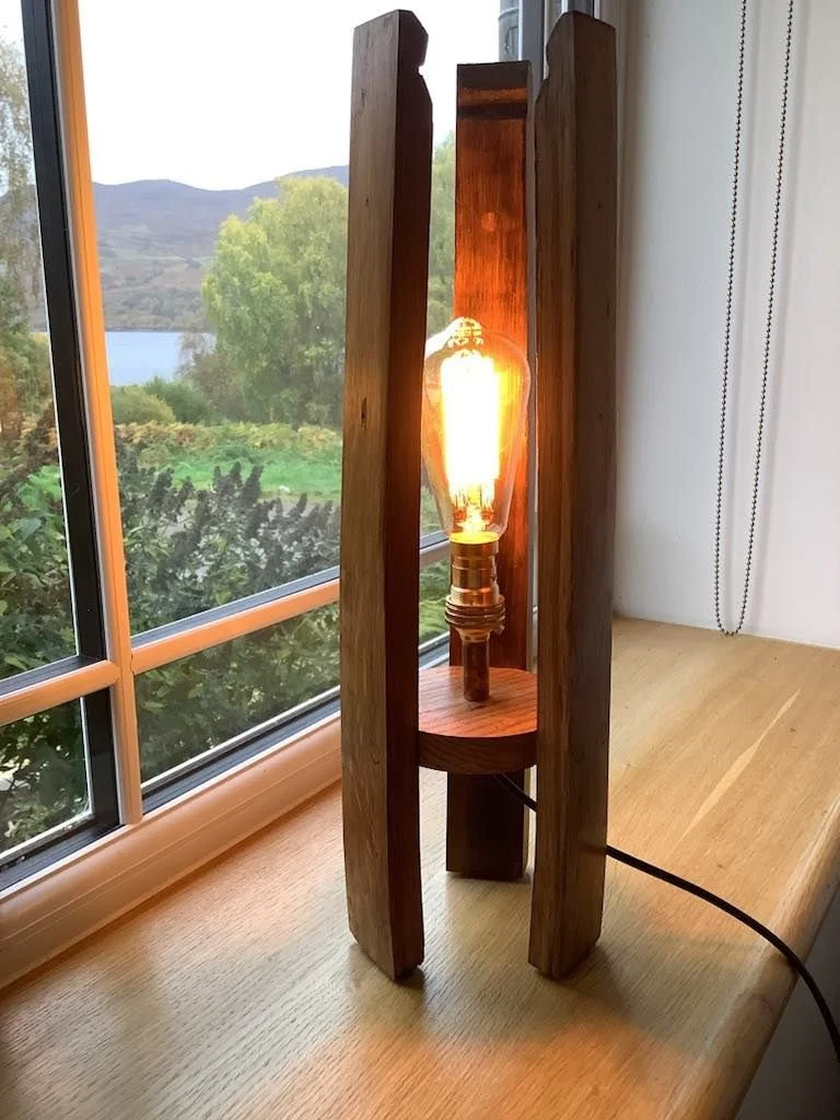 A wooden lamp with a metal light bulb inside, placed on a wooden surface near a window with a landscape view of trees, hills, and a lake.