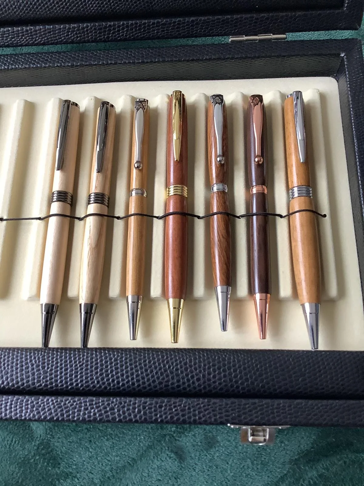 A set of seven wooden and metallic ballpoint pens in a case, arranged in two rows with a black elastic band holding them in place.