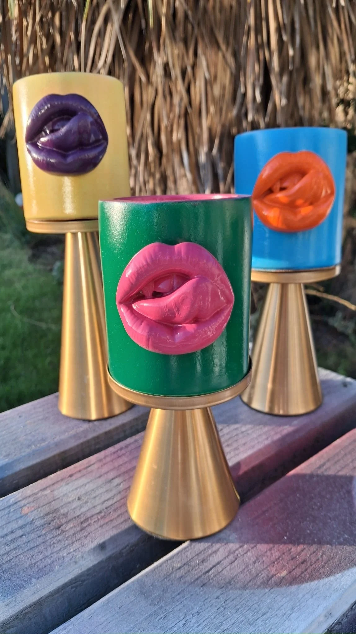 Colorful lamps with lips designs on top, each on a gold stand, outdoors with a wooden surface and foliage background.
