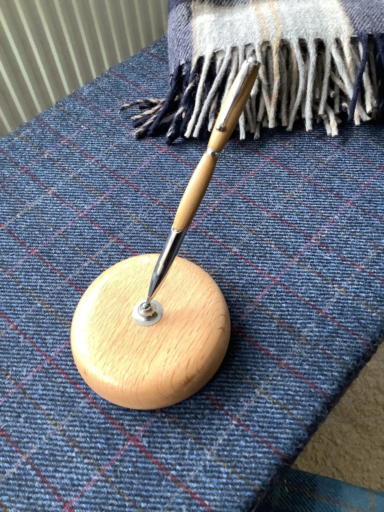 A wooden desk accessory holding a pen, placed on a blue checkered fabric surface, with a striped woven blanket in the background.