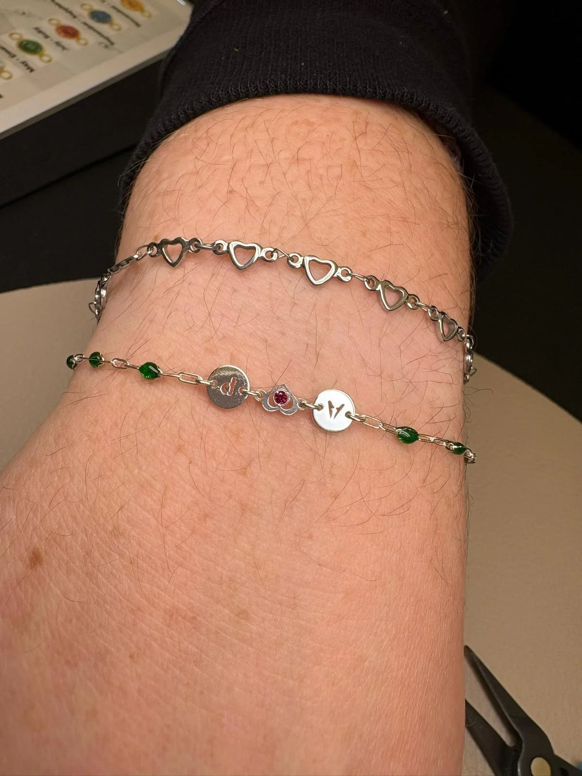 A person's arm with two bracelets: a silver heart chain bracelet and a silver chain bracelet with various charms and beads.