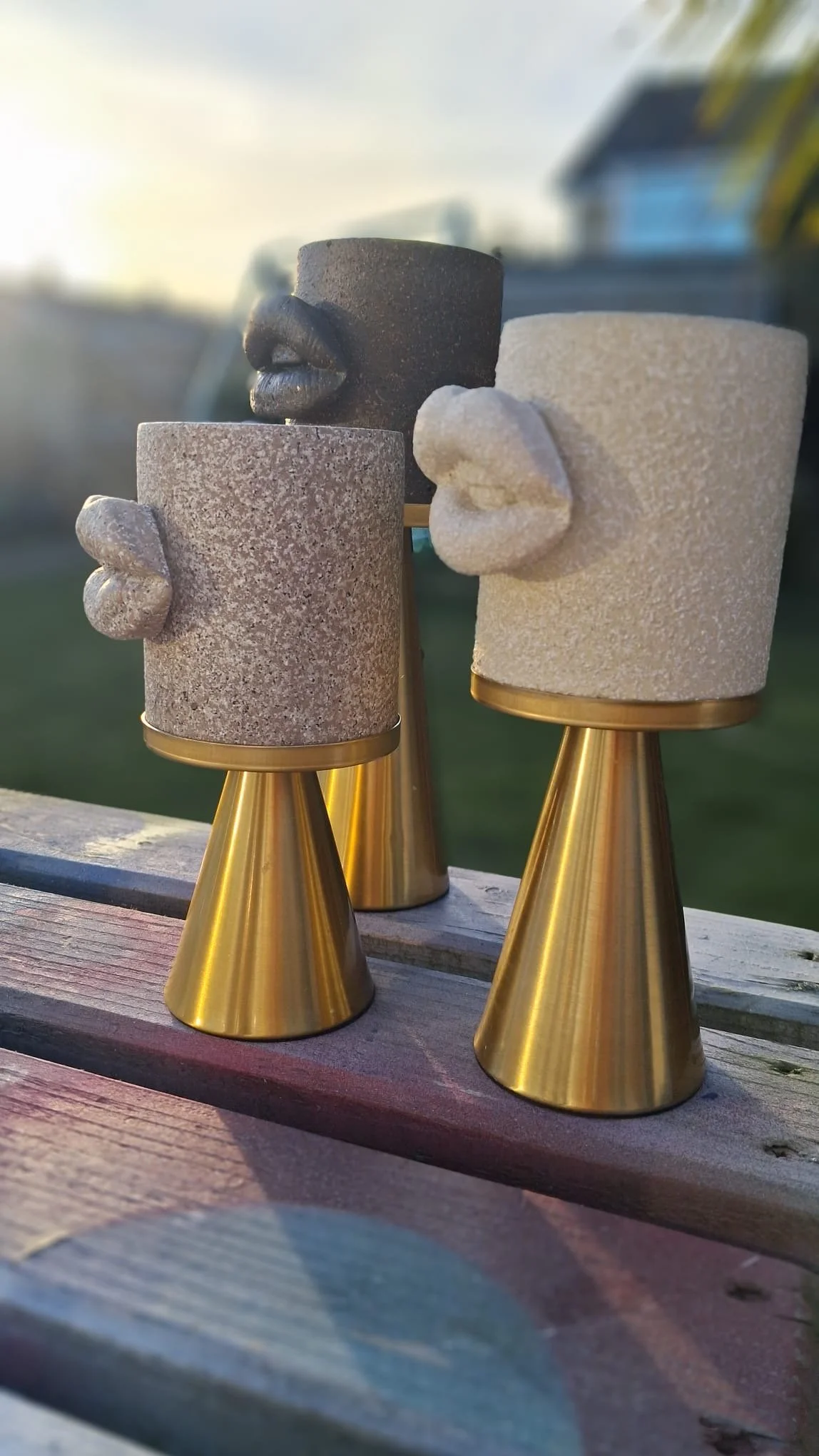 Decorative candle holders with abstract faces and lips, made of textured stone and glossy black and white material, placed on a wooden surface outdoors at sunset.
