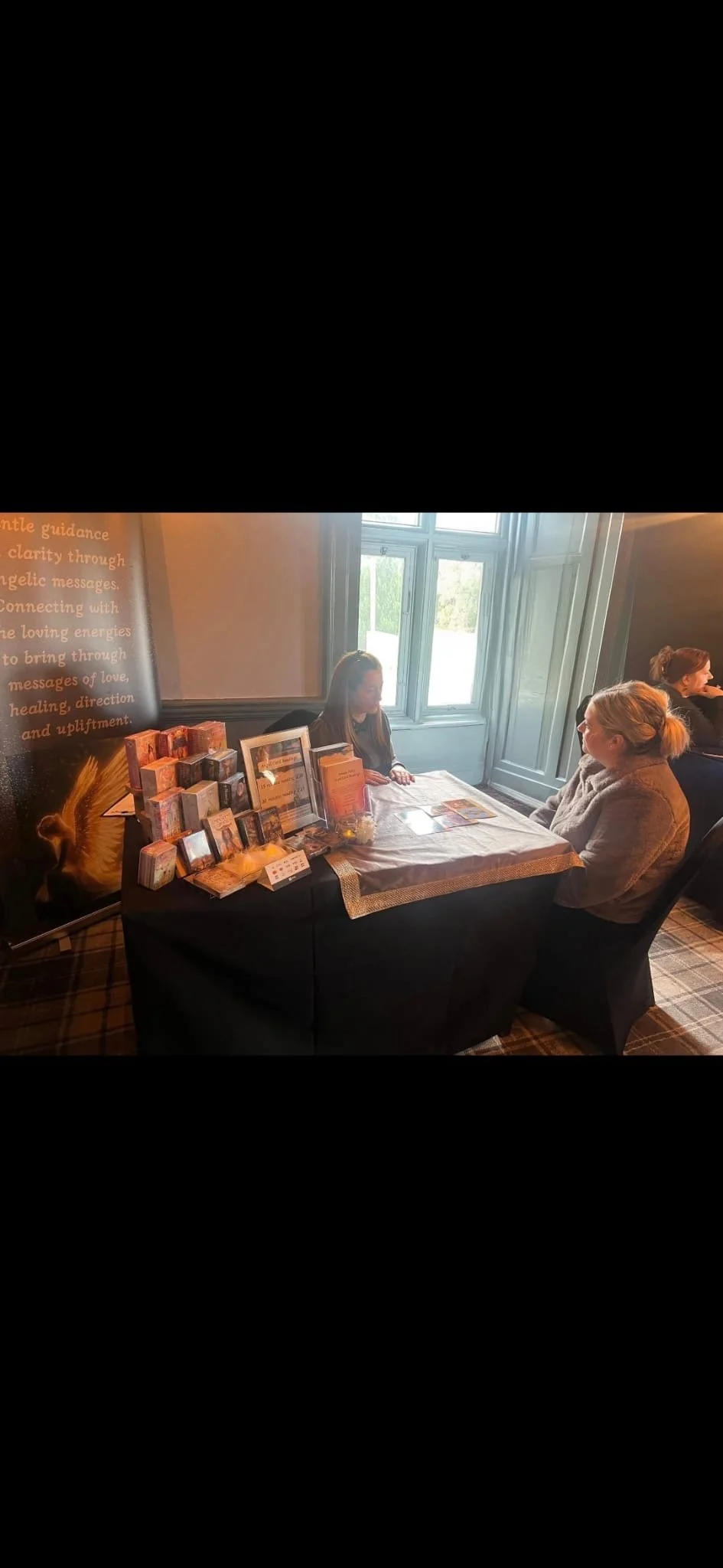 A woman sitting at a table with tarot cards, healing crystals, and spiritual books, talking to a woman on the other side of the table. The setup suggests a spiritual or tarot reading session in a well-lit room with large windows.