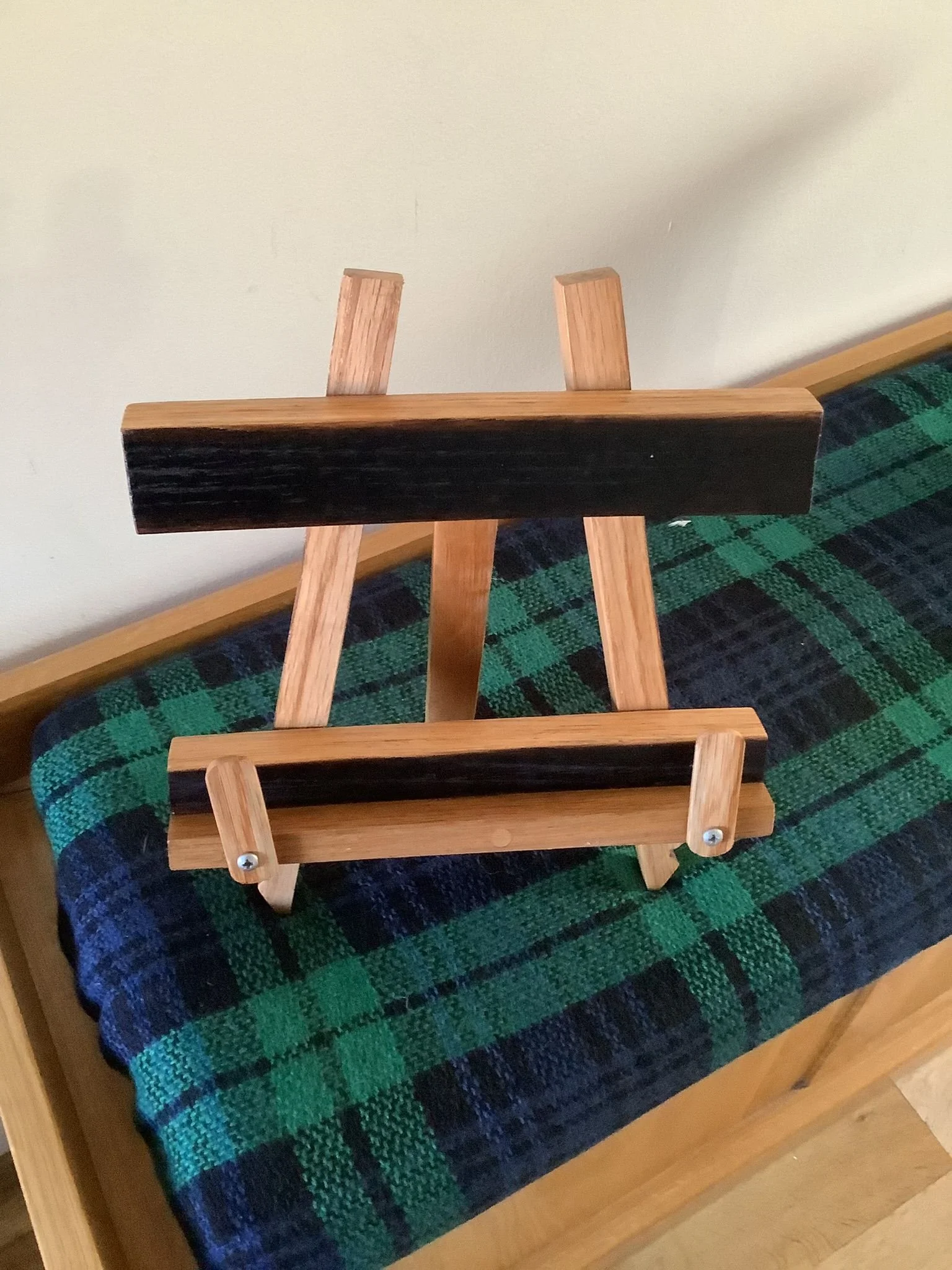 Small wooden easel standing on a plaid fabric surface, with a black painted bar across the top.