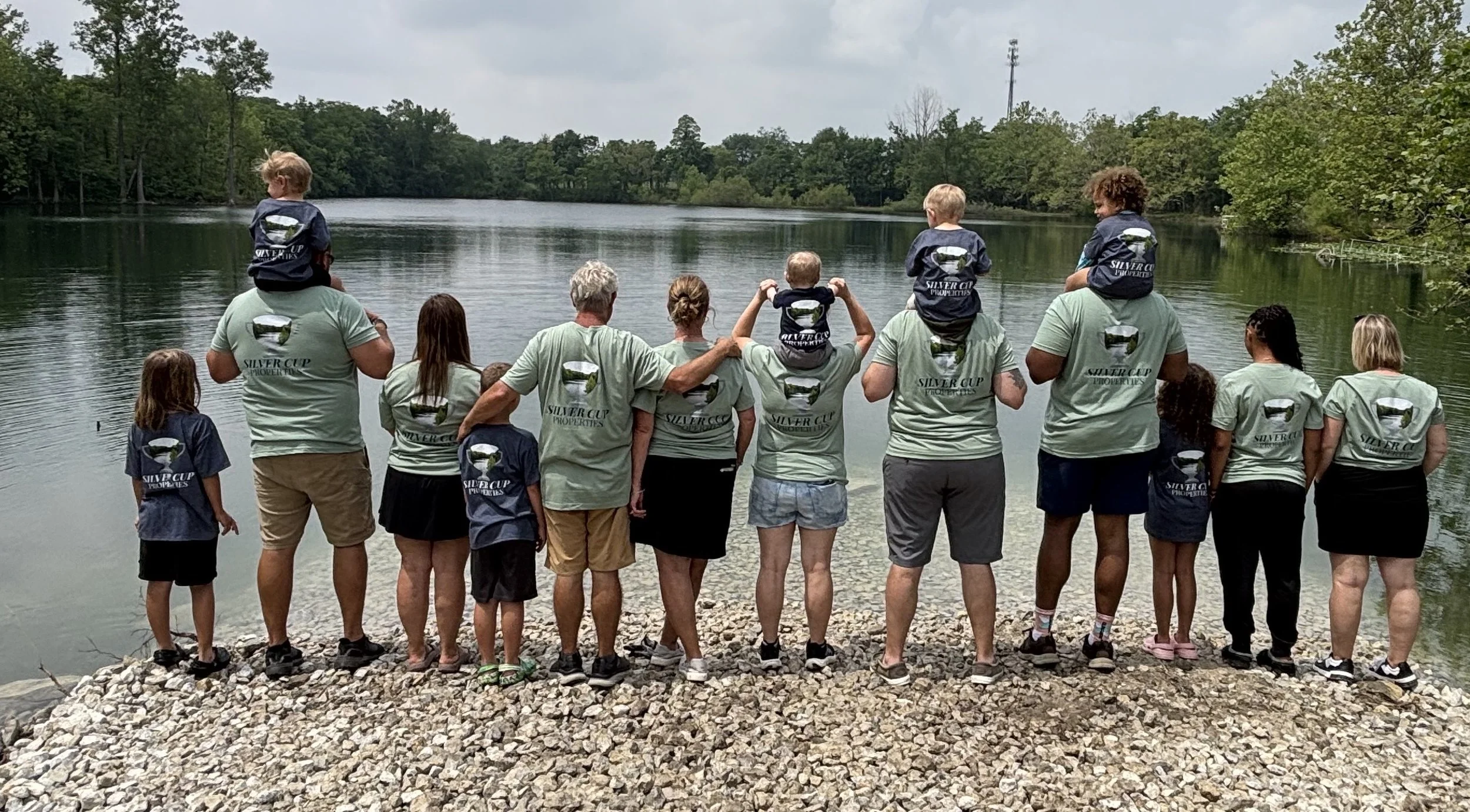 A group of children and adults standing on a rocky shoreline by a lake, wearing matching shirts with a whale logo, some children are sitting on adults' shoulders, and some are being carried.