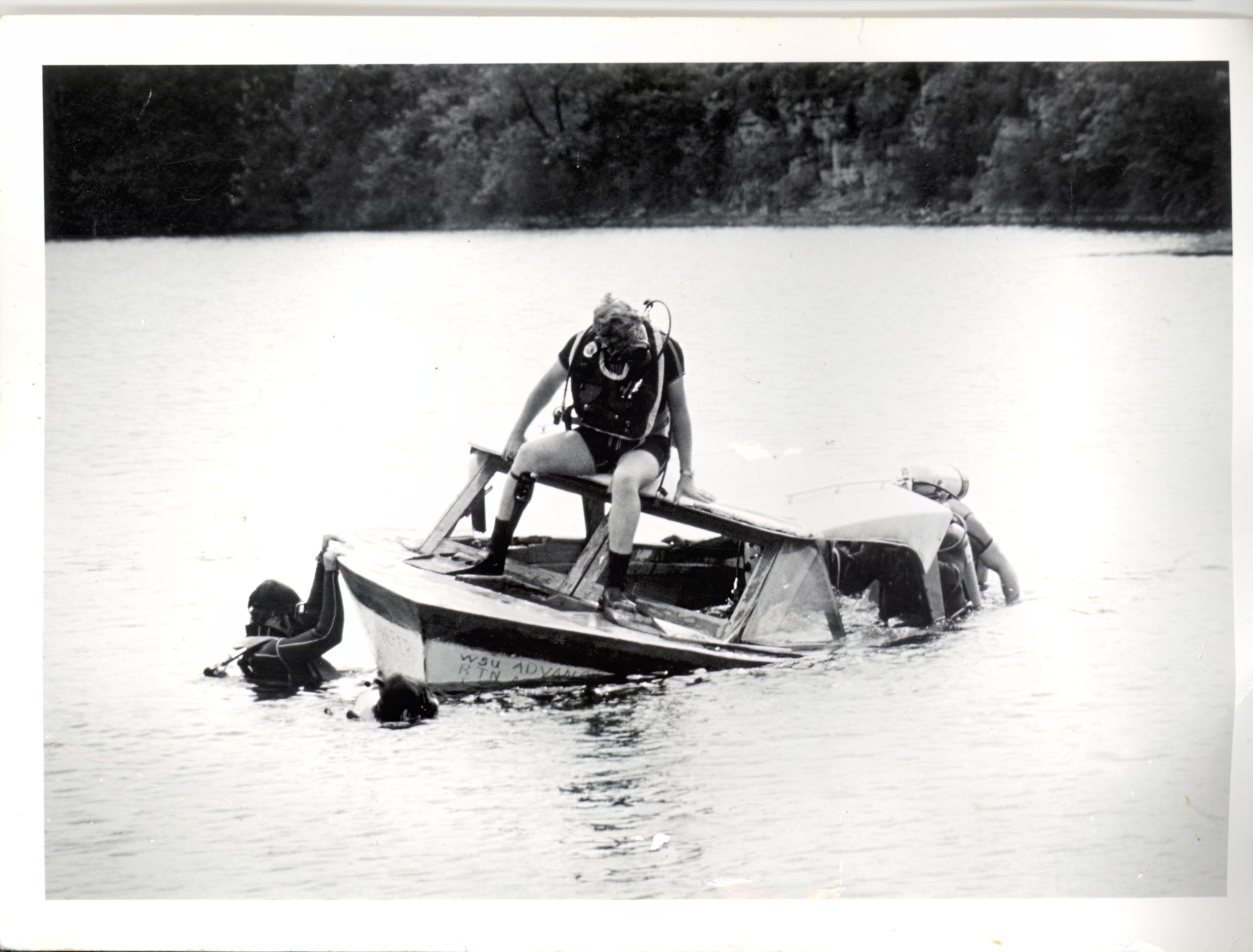Black and white photo of a person, wearing scuba gear, sitting on the edge of a partially submerged boat in a body of water, with two people in the water near the boat.