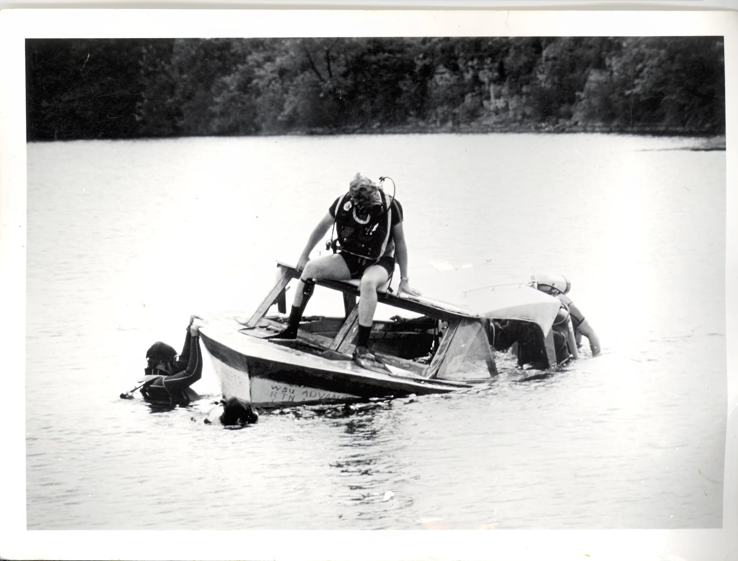 Black and white photo of a person, wearing scuba gear, sitting on the edge of a partially submerged boat in a body of water, with two people in the water near the boat.
