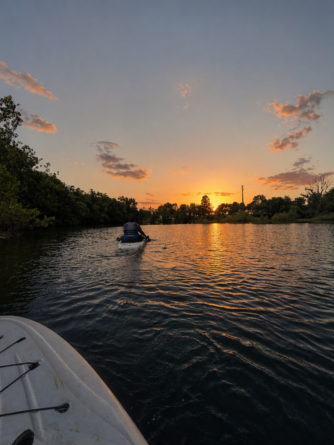 A person wearing a life jacket paddling a kayak on a calm river during sunset, with trees and a utility pole in the background. The photo is taken from another kayak or boat in the water.