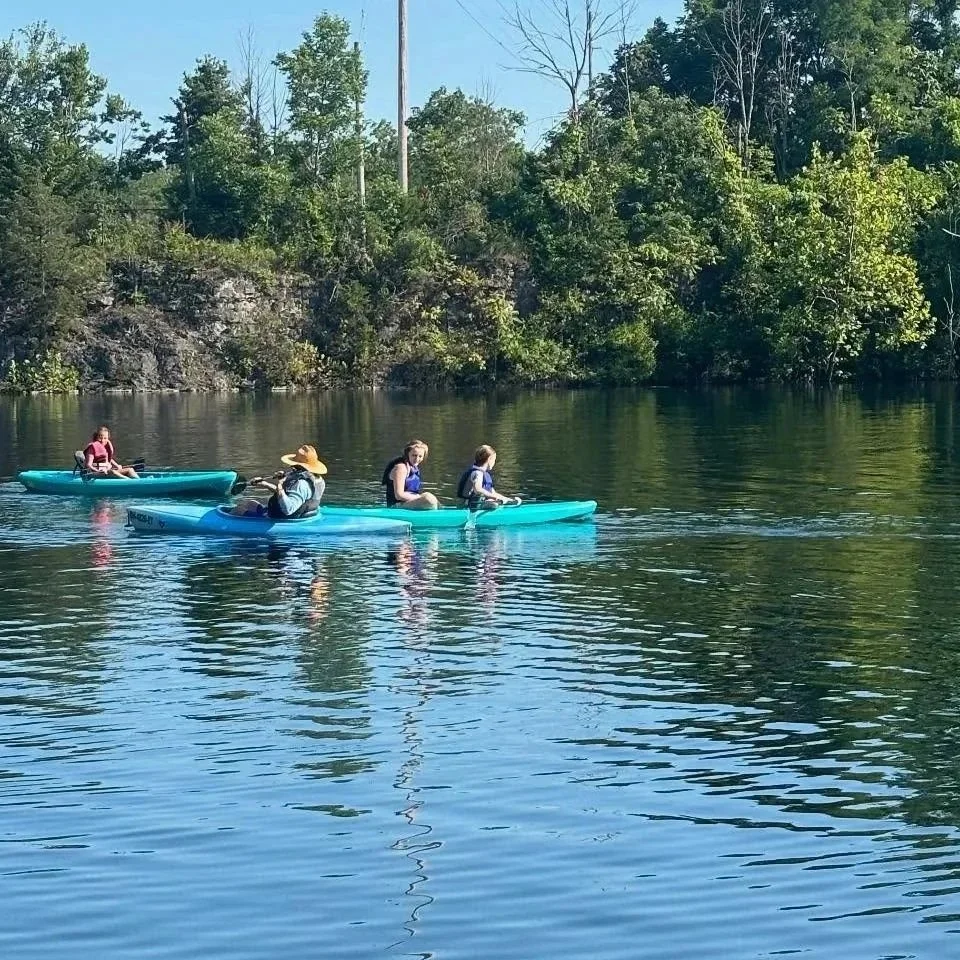 Four people kayaking on a calm river with trees and rocky banks in the background under a clear, sunny sky.