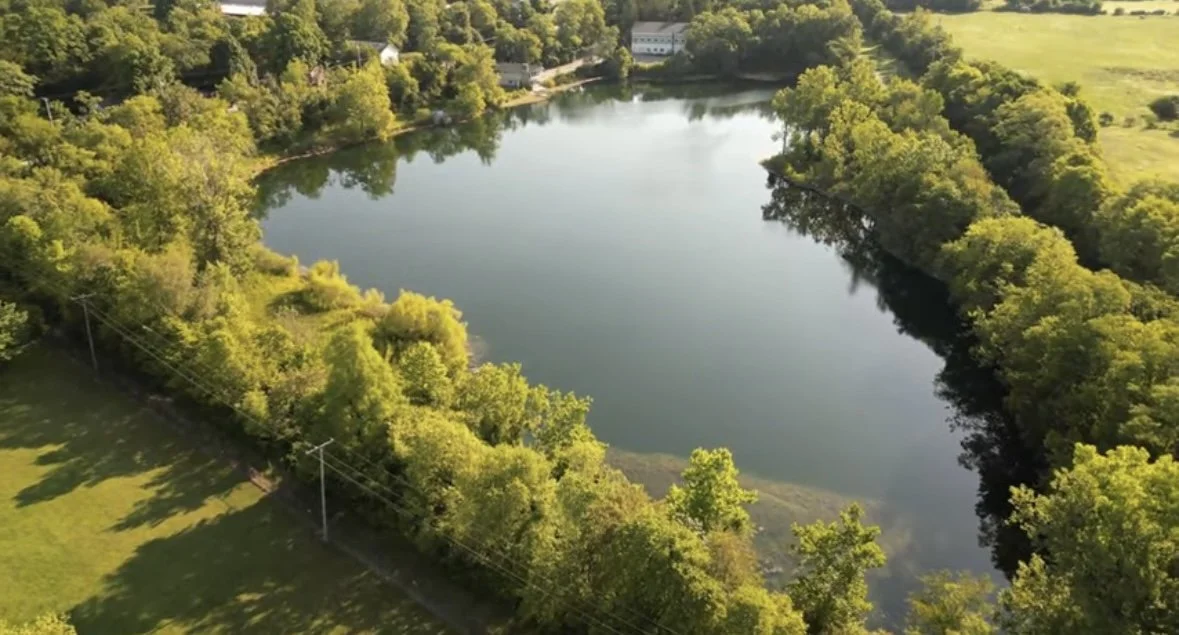 An aerial view of a small lake surrounded by dense green trees and open grassy areas.