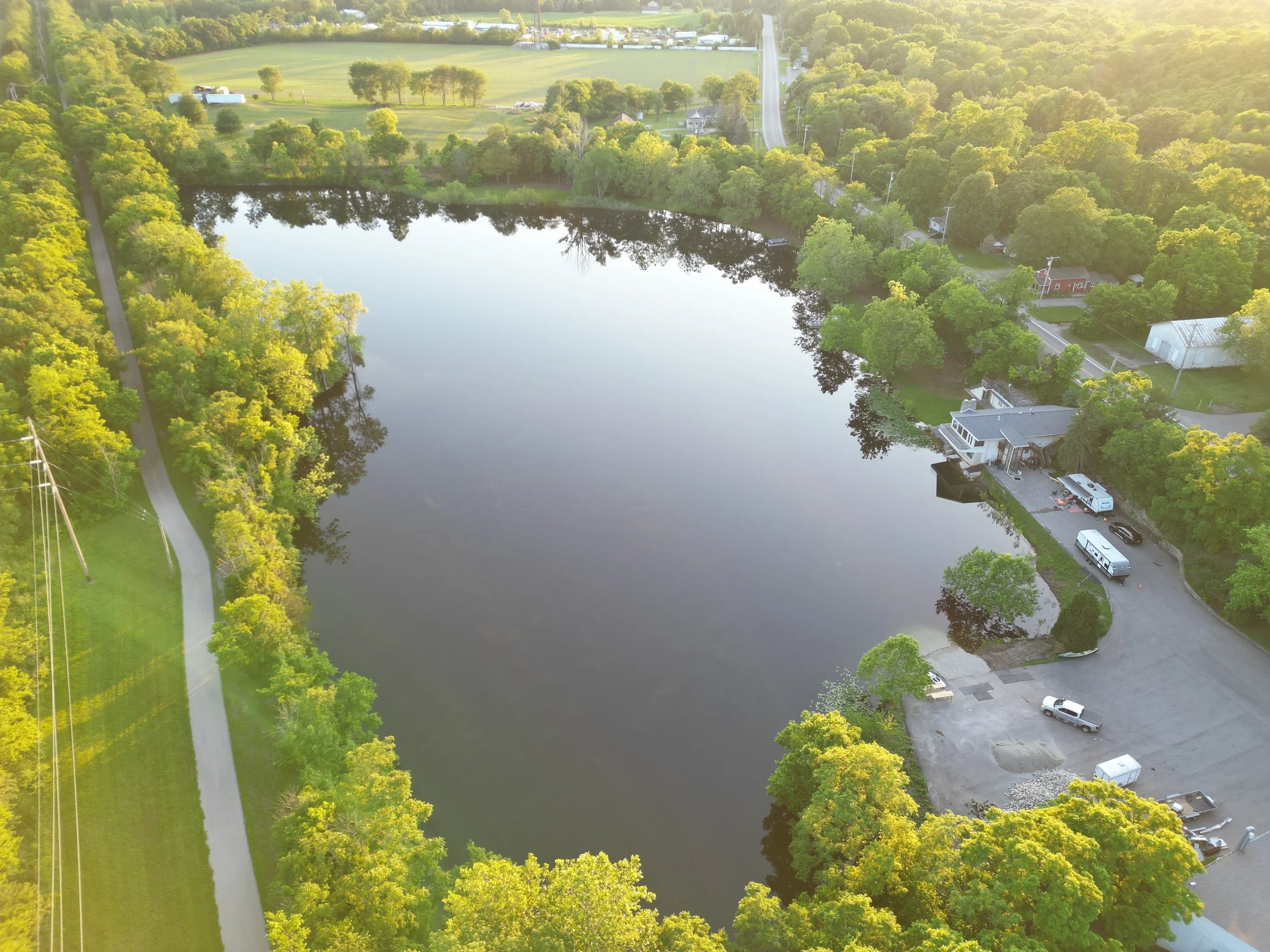 Aerial view of a small lake surrounded by greenery, houses, a parking lot with vehicles, and a road with trees along the side, during the daytime with bright sunlight.