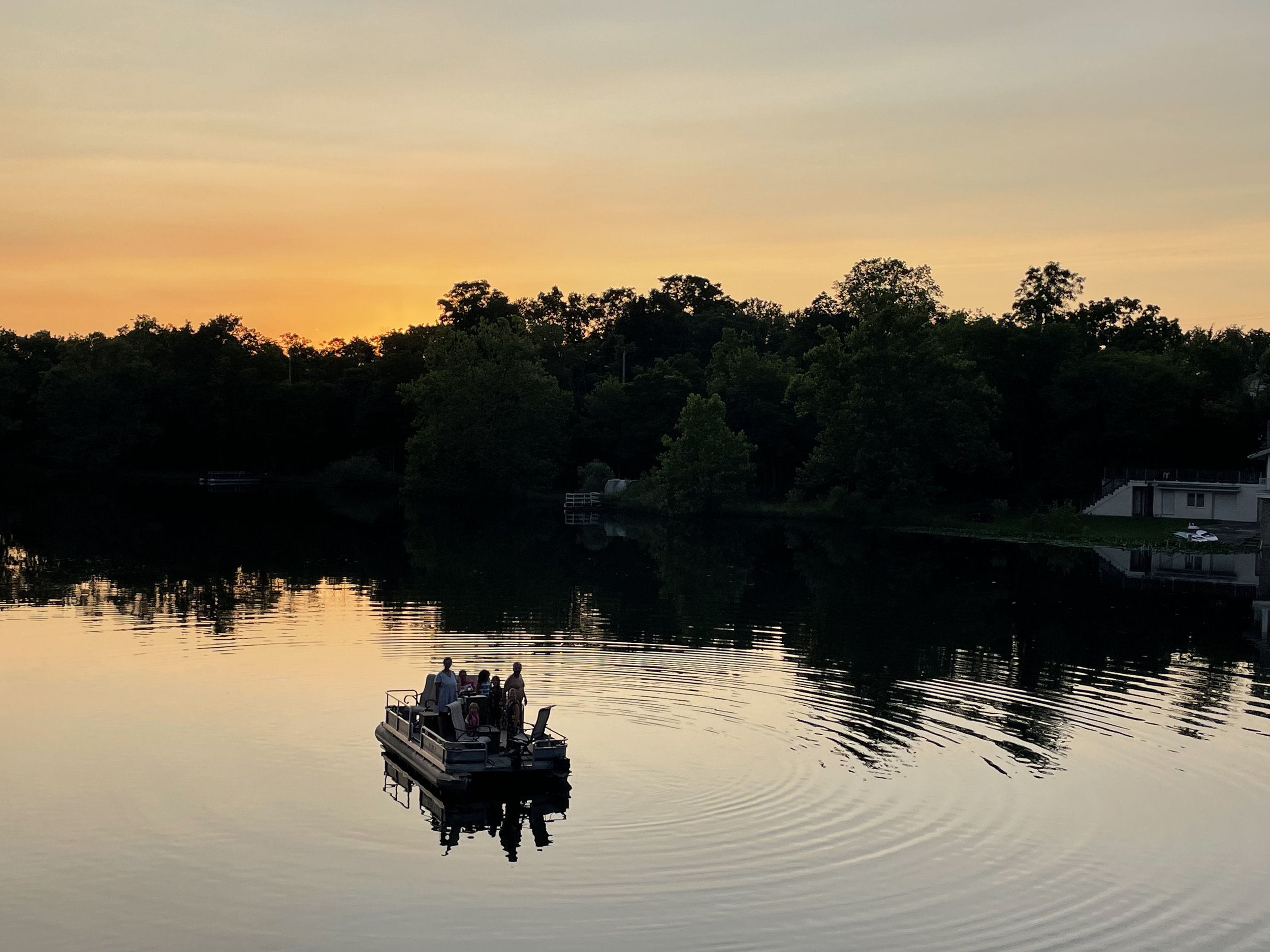 A small boat with several people aboard, sailing on a calm lake at sunset, with a wooded shoreline and houses in the background.