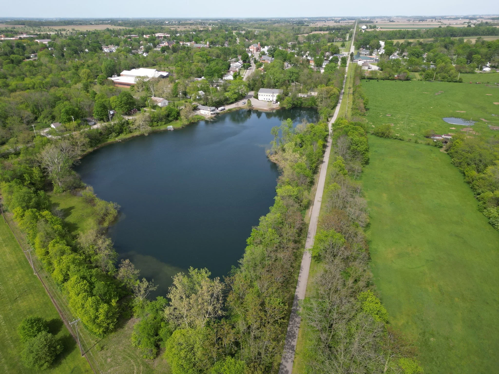 Aerial view of a small lake surrounded by trees, with a narrow walking path running parallel to a road on the right. Residential houses and green fields are visible in the background.