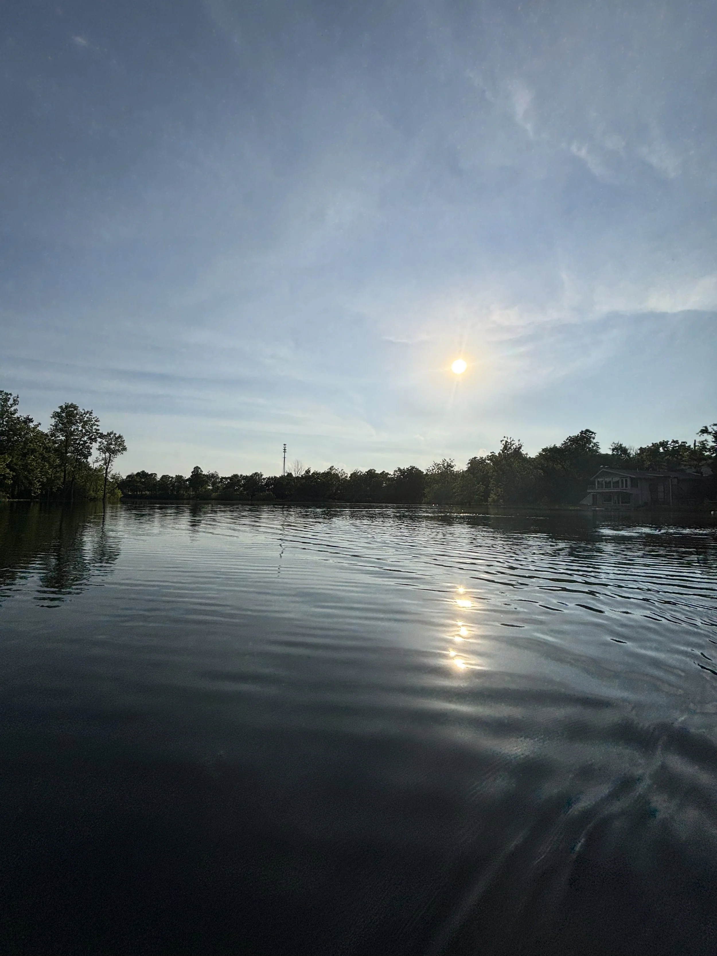 A serene lake reflecting the sun and partly cloudy sky, with trees and houses along the shoreline.