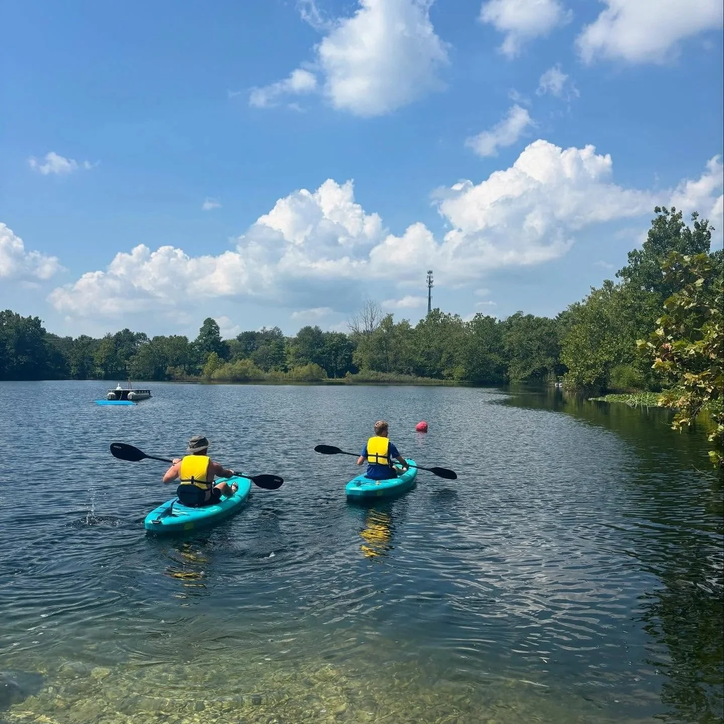 Two people in kayaks with yellow life jackets paddle on a calm lake under a partly cloudy sky, with trees along the shoreline and a boat in the distance.