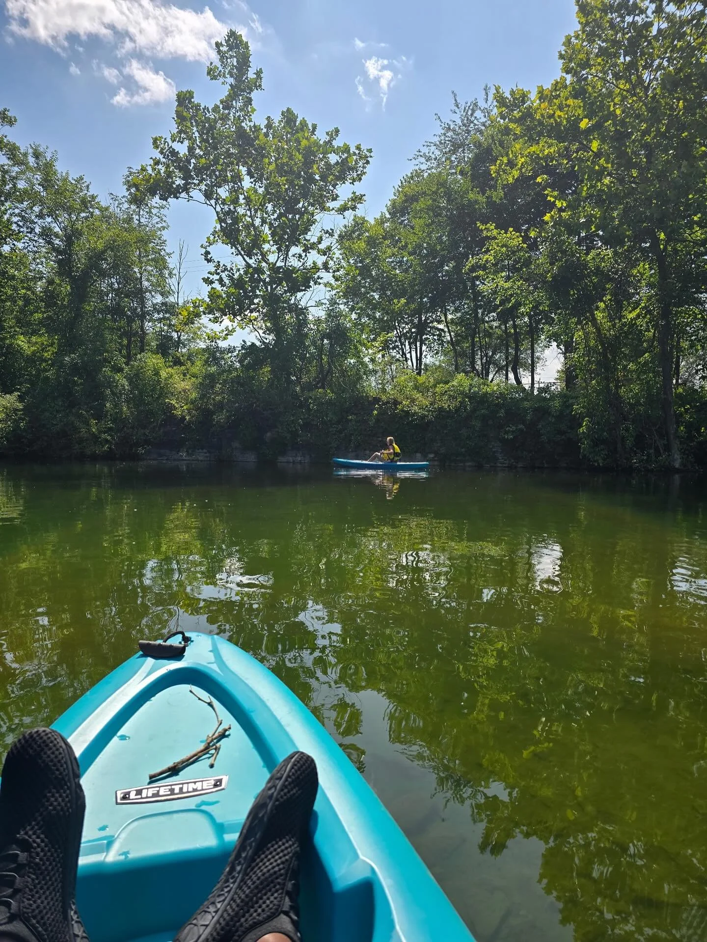 View from a kayak on a calm river with lush green trees on the riverbank and a person in a kayak in the distance.