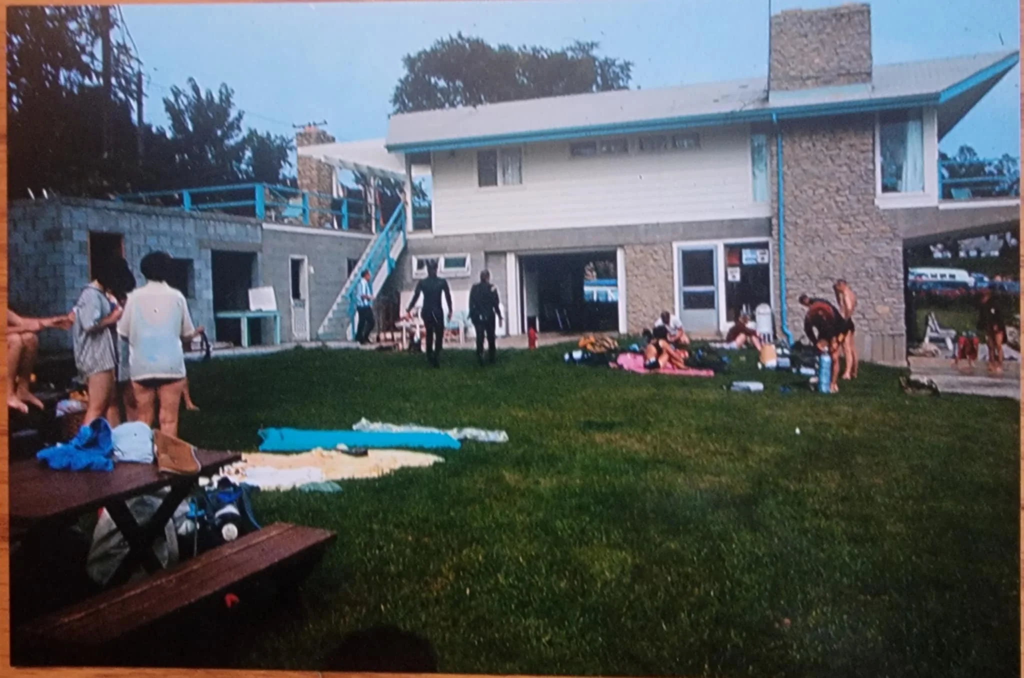 People gathered on a grassy lawn outside a two-story building what was once known as sportsmans lake with stone and wooden siding, some sitting on blankets, with trees having fun