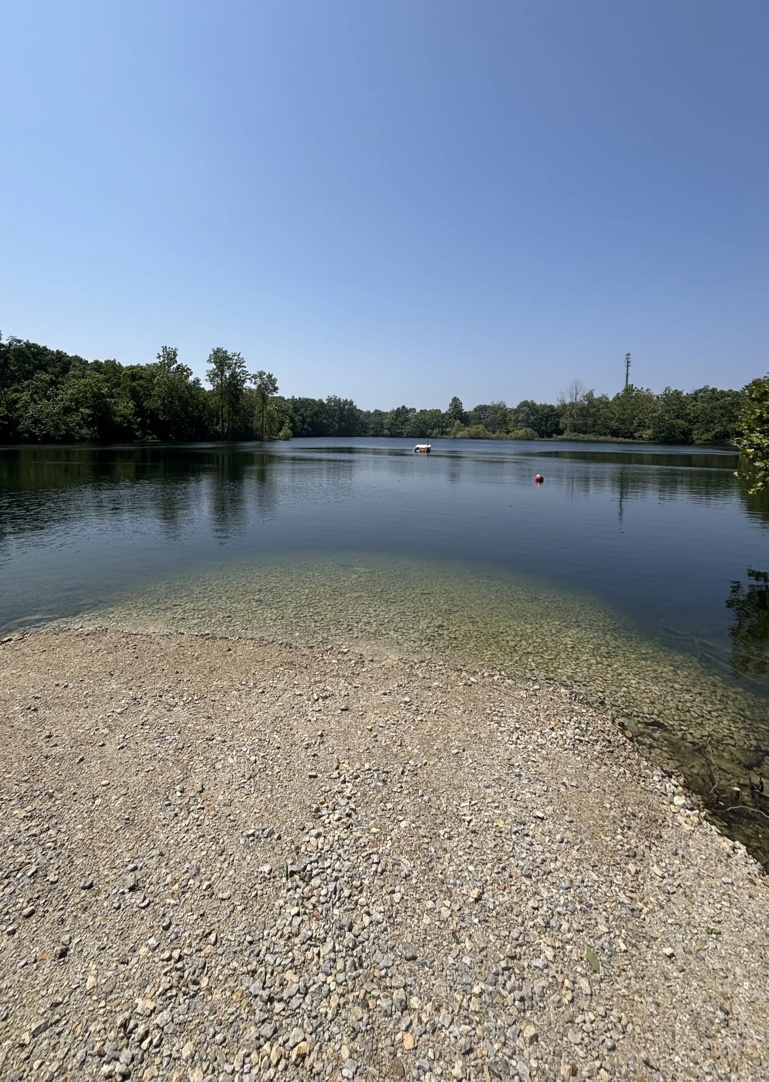 A calm river with a boat floating on the water, surrounded by green trees under a clear blue sky, with a gravelly shoreline in the foreground.