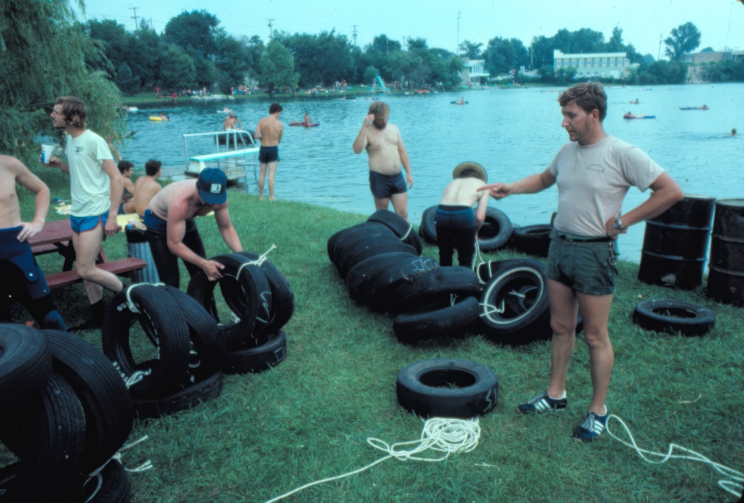 People by a lake, some sitting on tire stacks, some setting up tires for a boat race, others in swimsuits and shorts near the water on a grassy bank, with trees and buildings in the background.