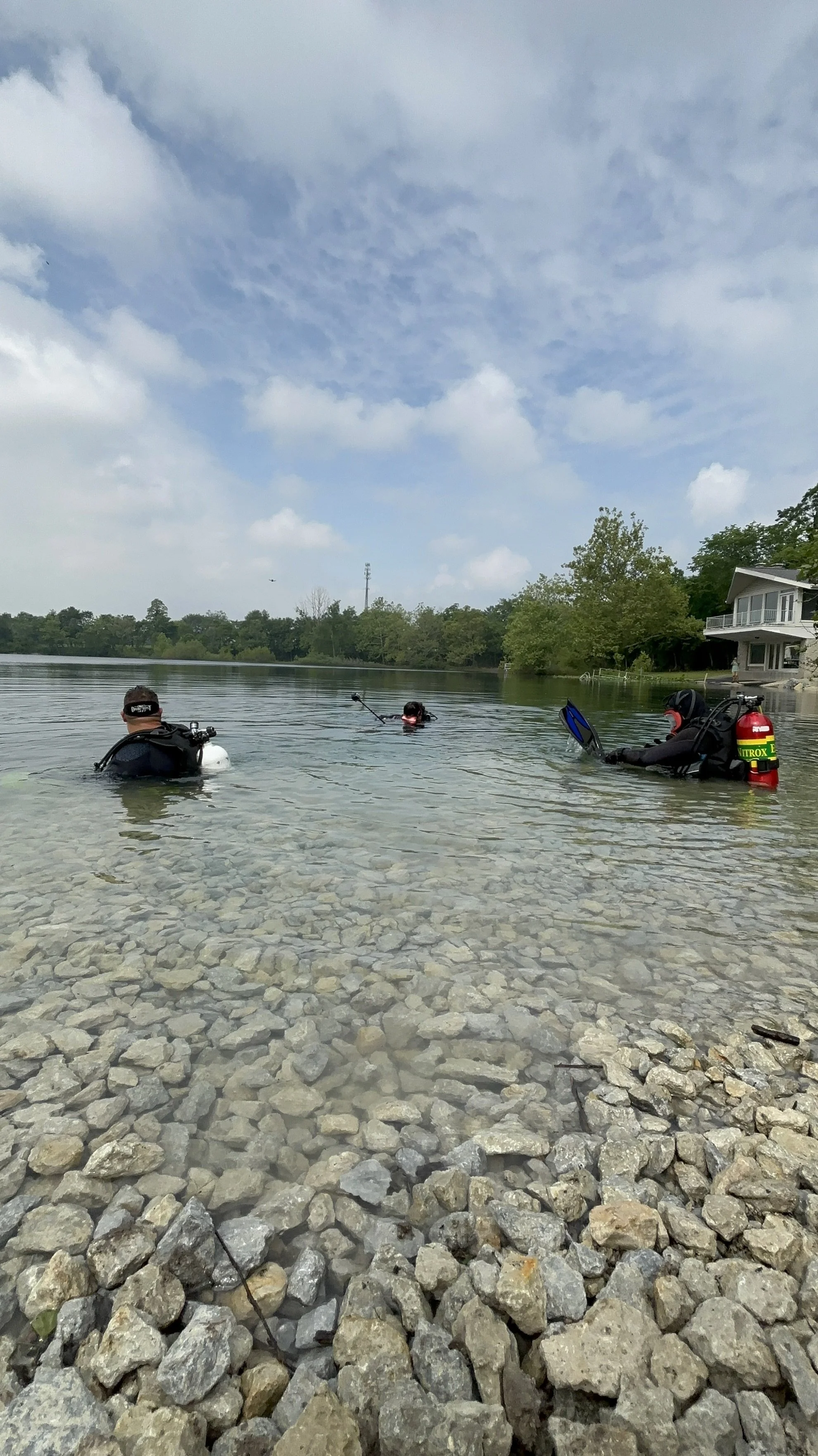 Three scuba divers in shallow water by a rocky shore, preparing for a dive with gear, near a lake with trees and a house in the background.