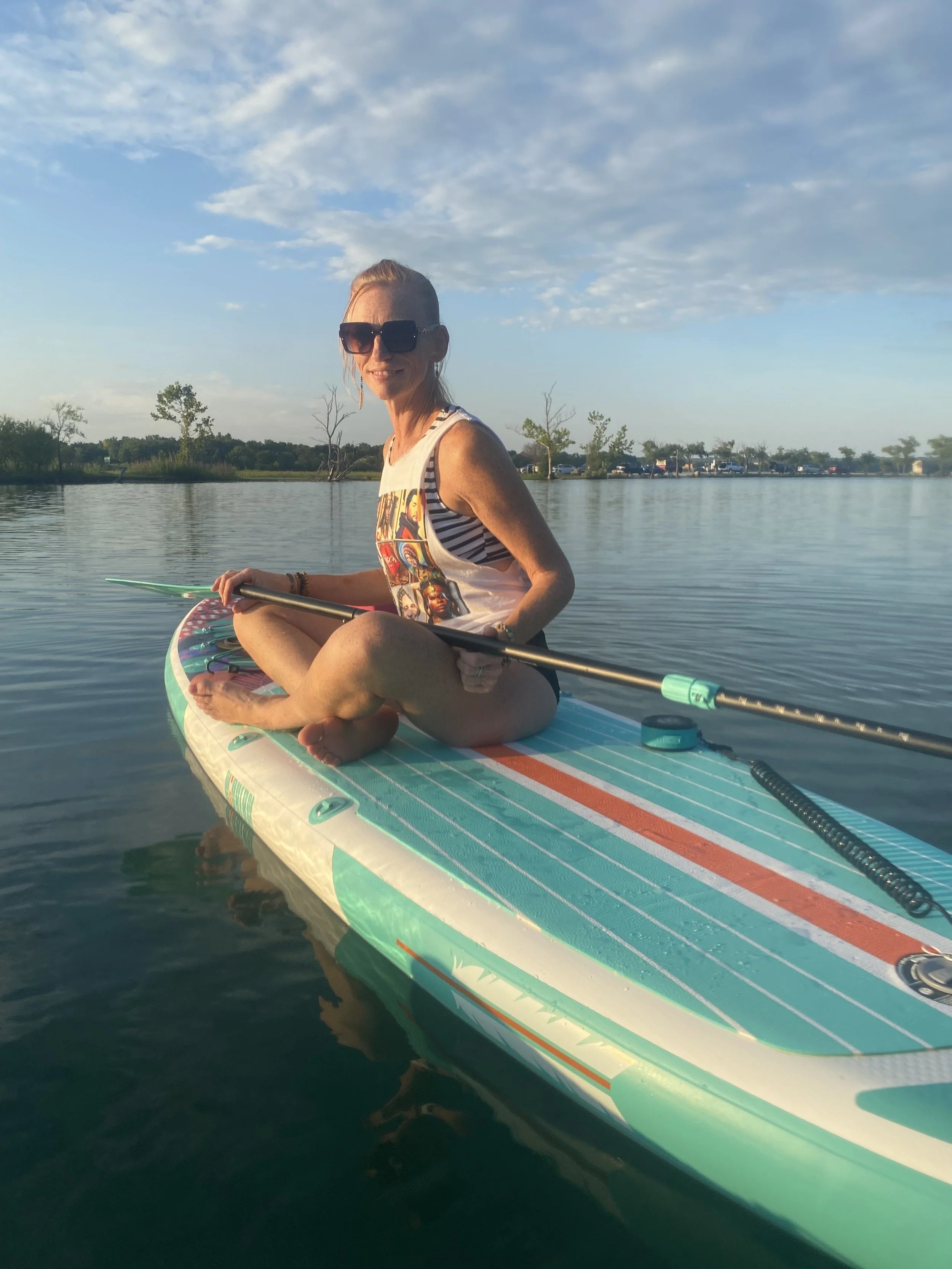 A woman with blonde hair, wearing large sunglasses and a colorful tank top, is sitting cross-legged on a paddleboard on calm water, holding a paddle, during a clear day with blue skies and scattered clouds.