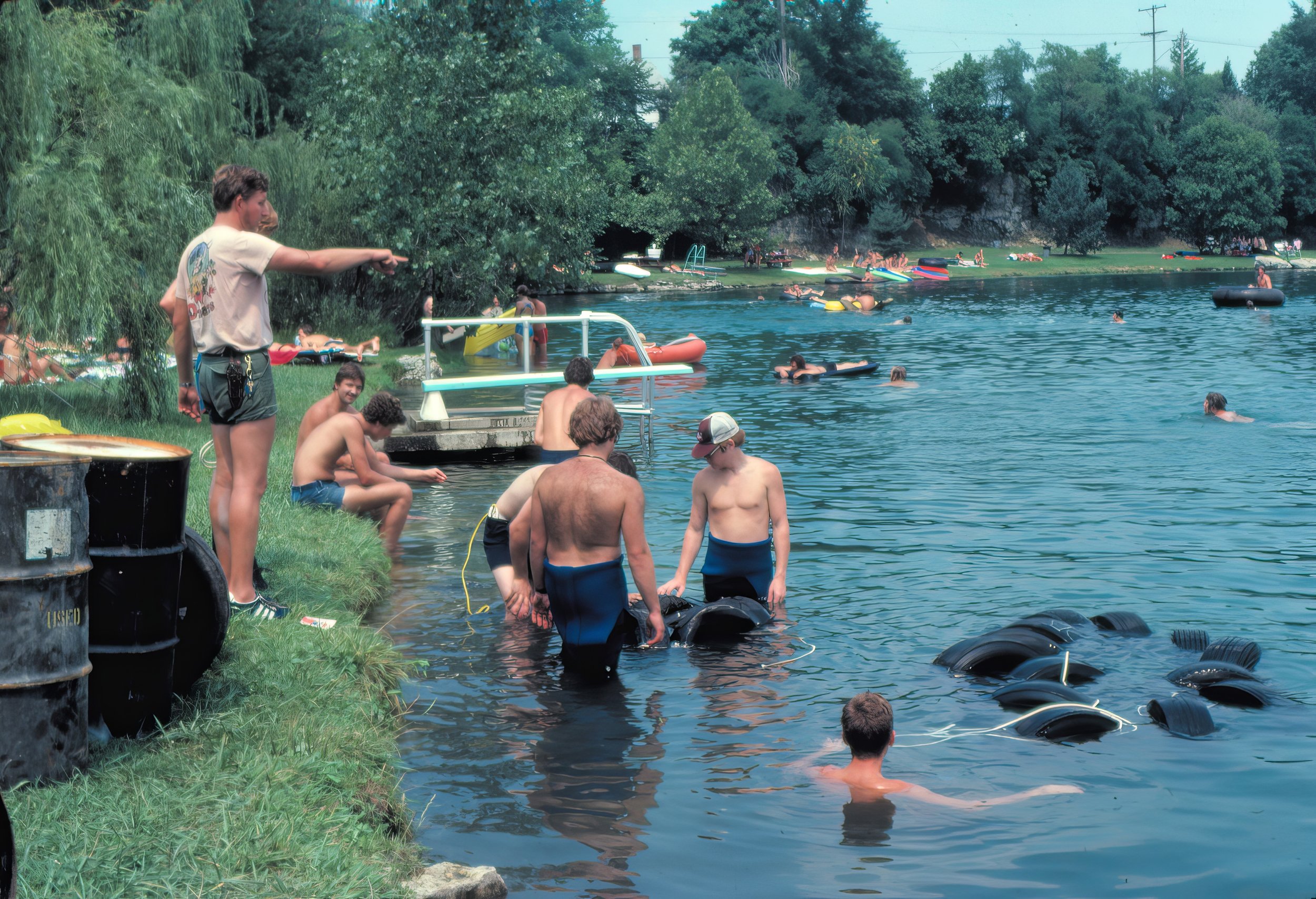 People swimming and relaxing by a lakeside with trees, boats, and floatation devices in the background.