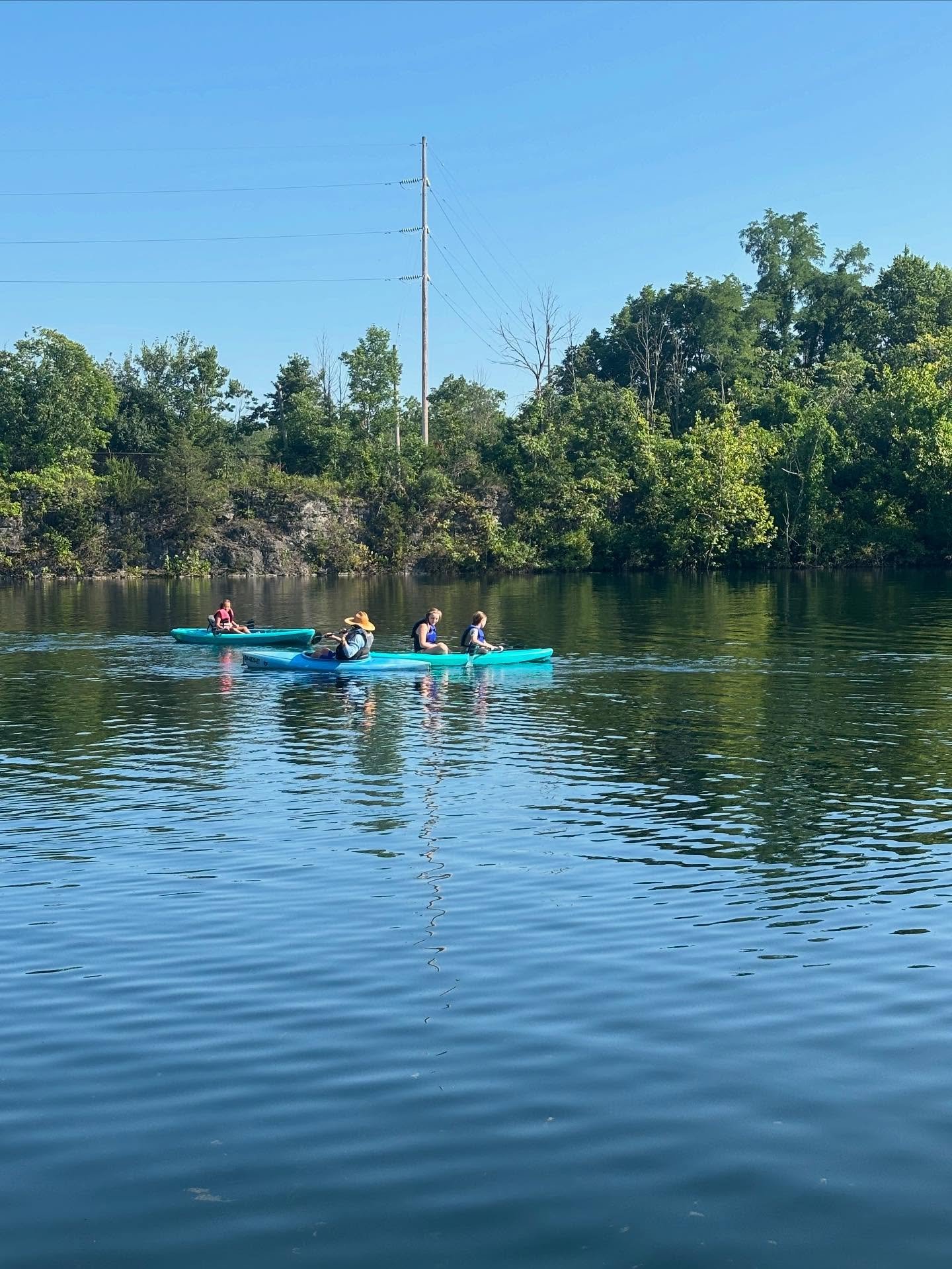Four people kayaking on a calm river, surrounded by trees and a clear blue sky.