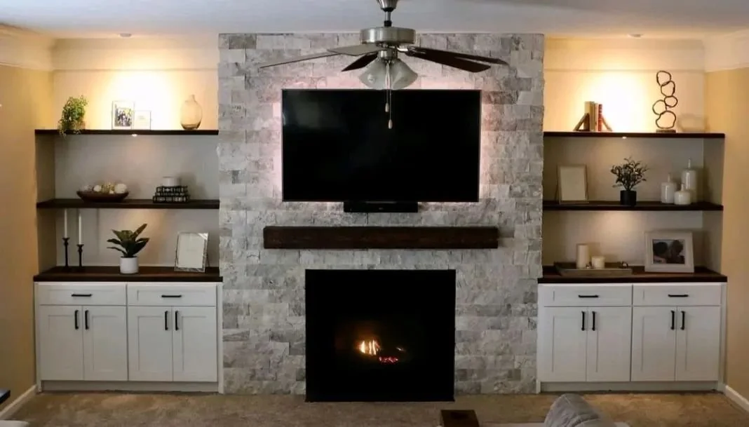 Living room with a stone accent wall, fireplace, and mounted TV above the fireplace. Two built-in shelves on each side with decorative items, candles, and plants. Ceiling fan with lights above.