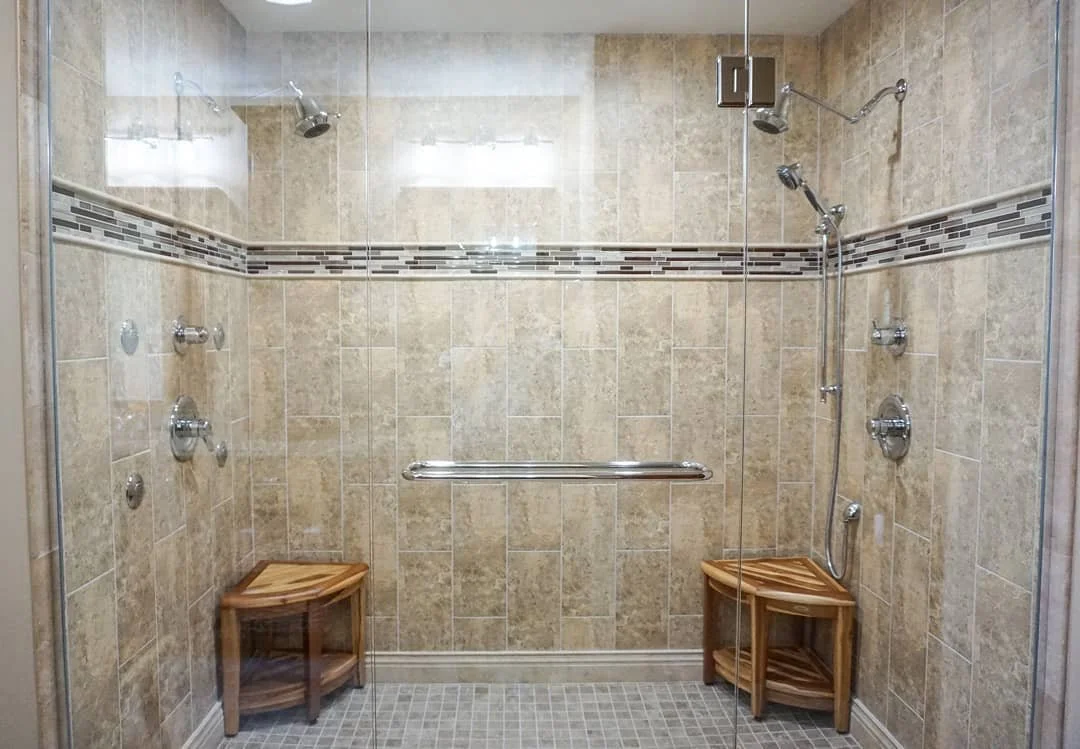 A spacious walk-in shower with beige tile walls, a horizontal decorative tile strip, multiple showerheads, and two small wooden stools.