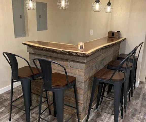 Wooden bar with four black and brown chairs around it, hanging pendant lights above, and a small box on the countertop.