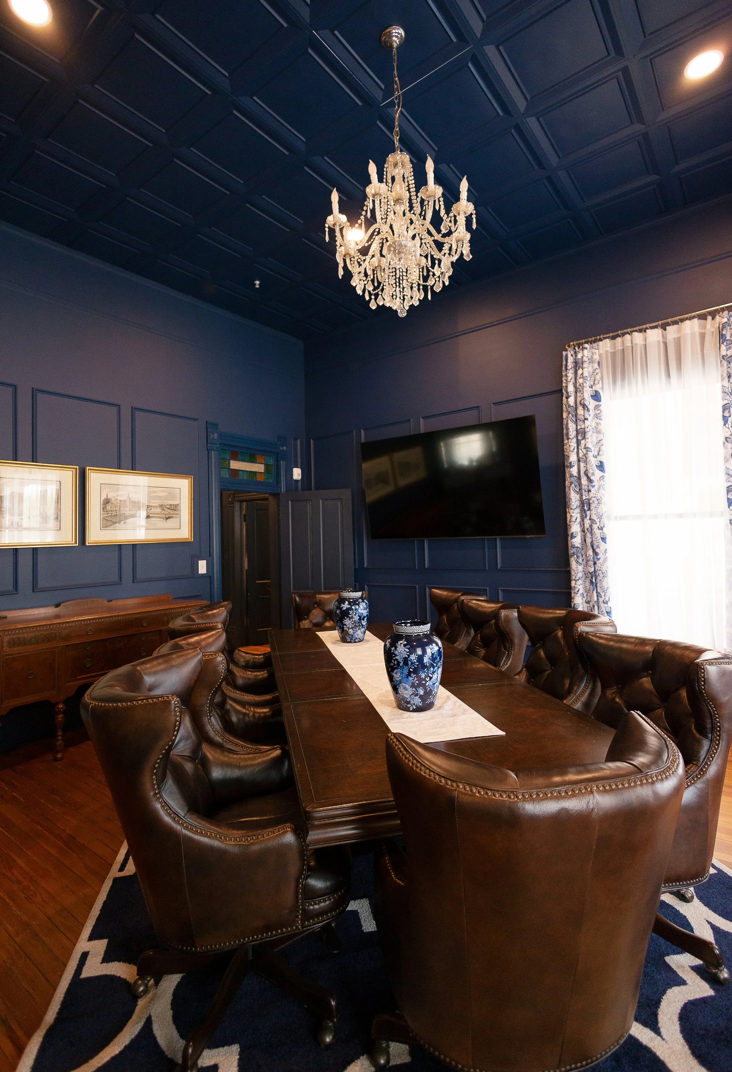 A formal dining room with a large dark wooden table surrounded by ten leather chairs. The table has three blue and white vases and a beige table runner. The room has dark blue walls with decorative paneling, a chandelier hanging from the ceiling, a large flat-screen TV mounted on the wall, and a window with patterned blue and white curtains.