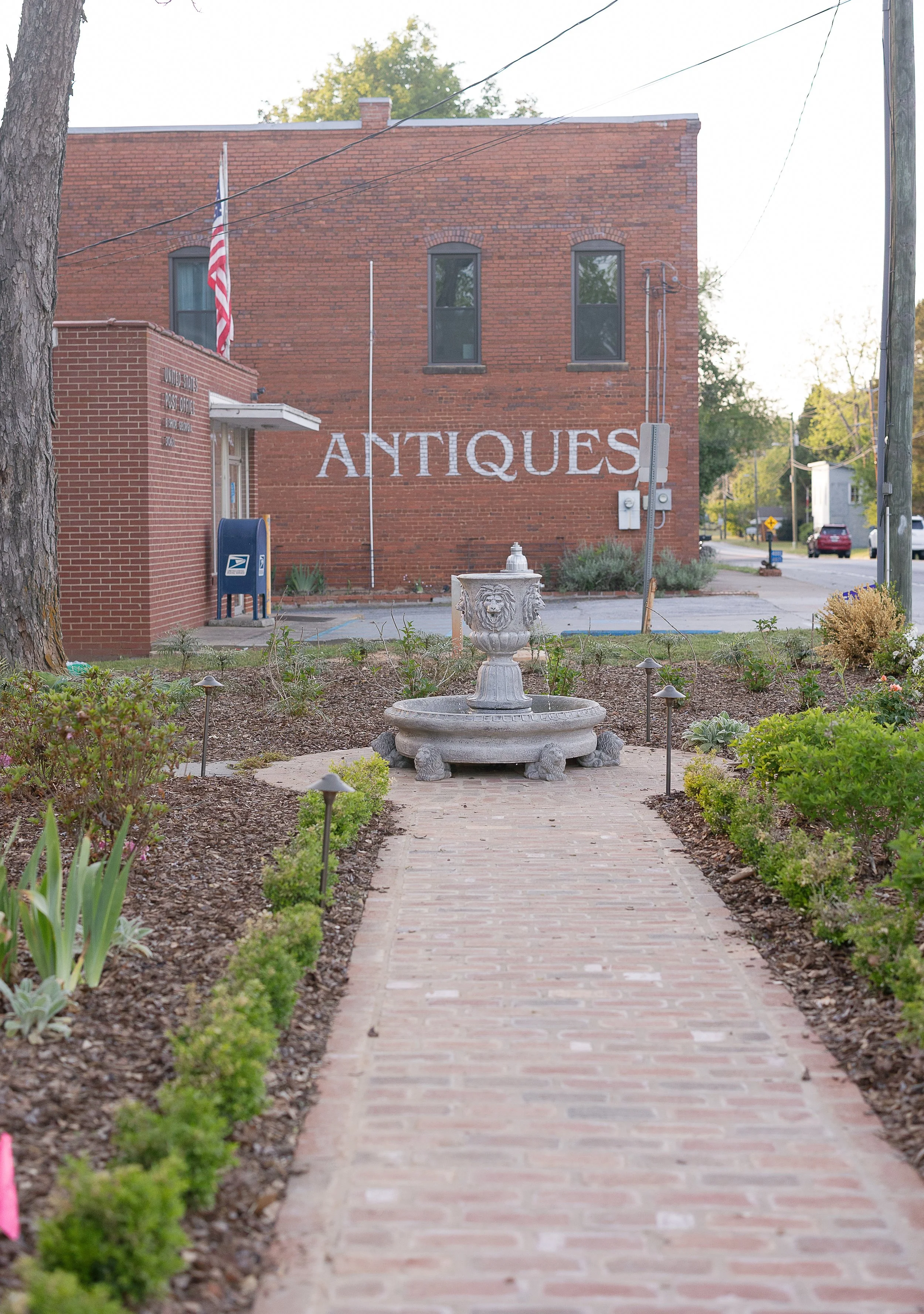 A sidewalk with brick paving leading towards a red brick building with a large white sign that says "ANTIQUES". There is a small fountain with a lion head sculpture in the center of the garden area along the pathway. The building has a small post office sign and American flag on the left side. There are plants and flowers lining the sidewalk on both sides, and trees and cars are visible in the background.