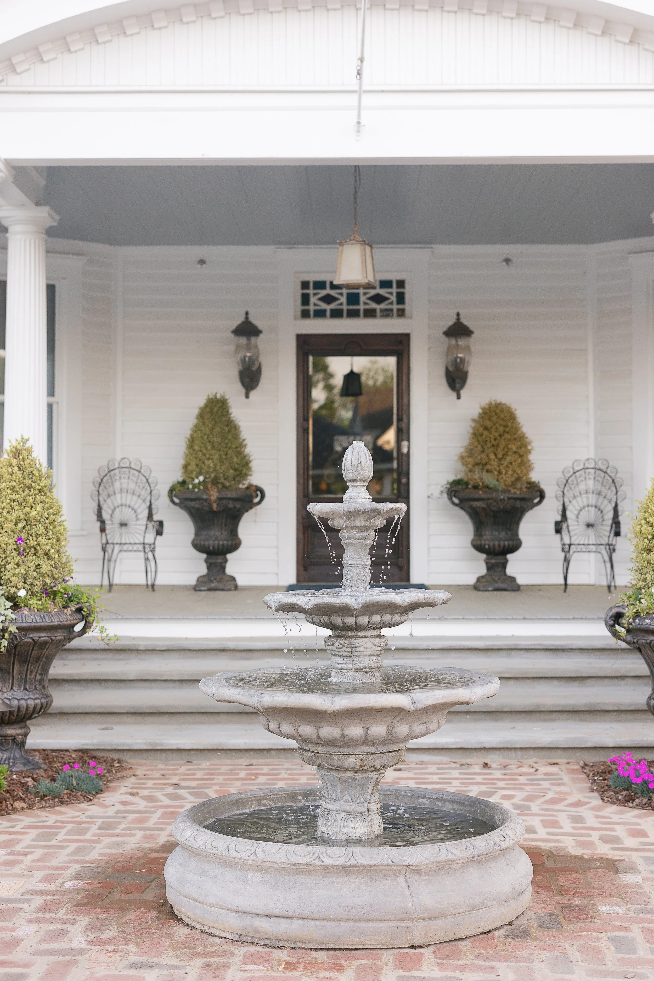 Front porch with a tiered fountain in the center, two potted shrubs on either side, black wrought iron chairs, and a wooden door with glass panels surrounded by white siding. Hanging lanterns are on either side of the door.