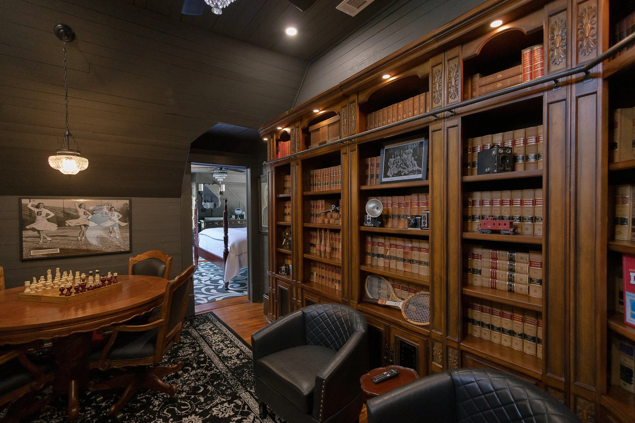 A cozy living room featuring a large wooden bookshelf filled with law books, a chess table with chairs, and a partially visible bed in a nearby room, with dark wood paneling on the ceiling and a decorative rug on the floor.