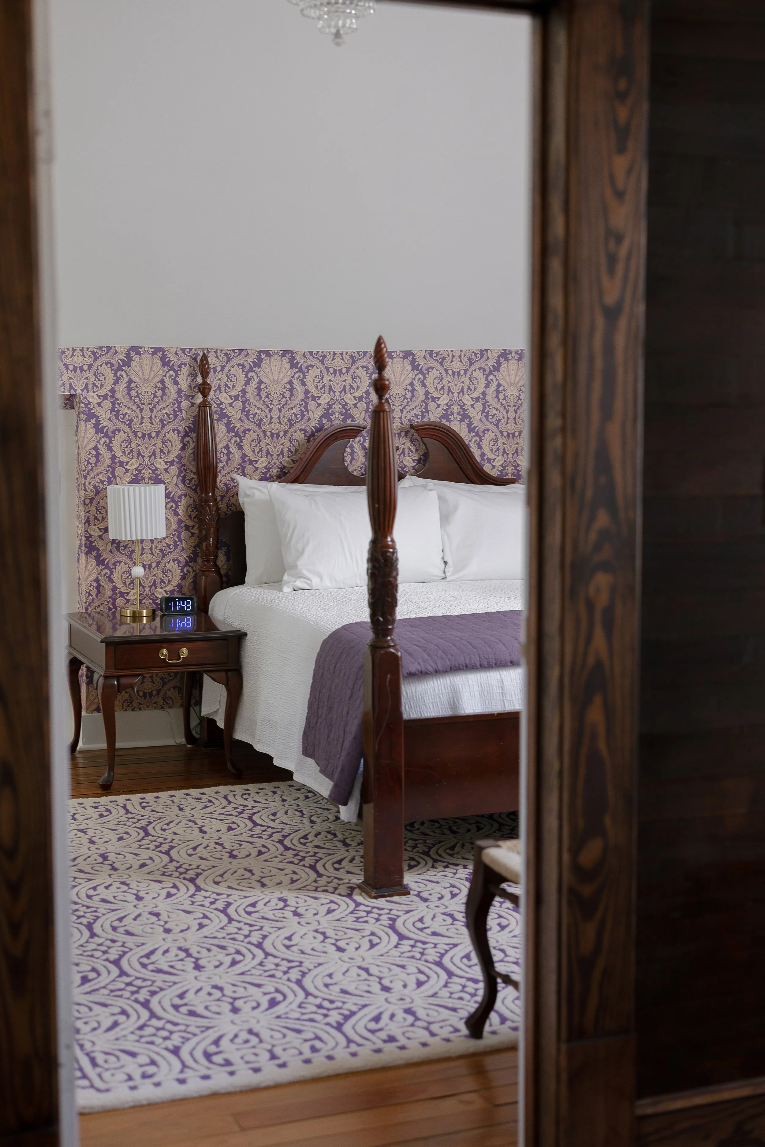 A bedroom with a four-poster bed, purple and white patterned bedding, and a matching purple and white patterned rug. The headboard is made of wood, and there is a nightstand with a digital clock and a white lamp. The room features purple and gold wallpaper above the bed, with white walls and a chandelier hanging on the ceiling. The photo is taken through a mirror frame.