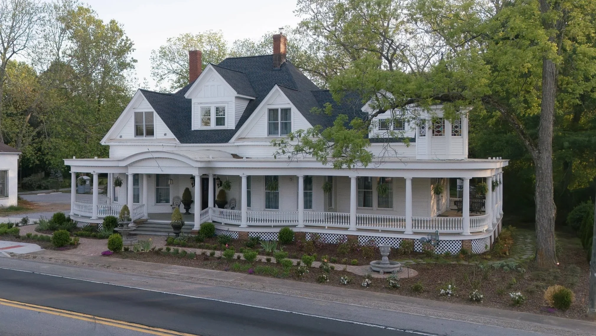 A large white Victorian-style house with a wrap-around porch, multiple gabled roofs, and a garden in front. The house is situated next to a street with a double yellow line.
