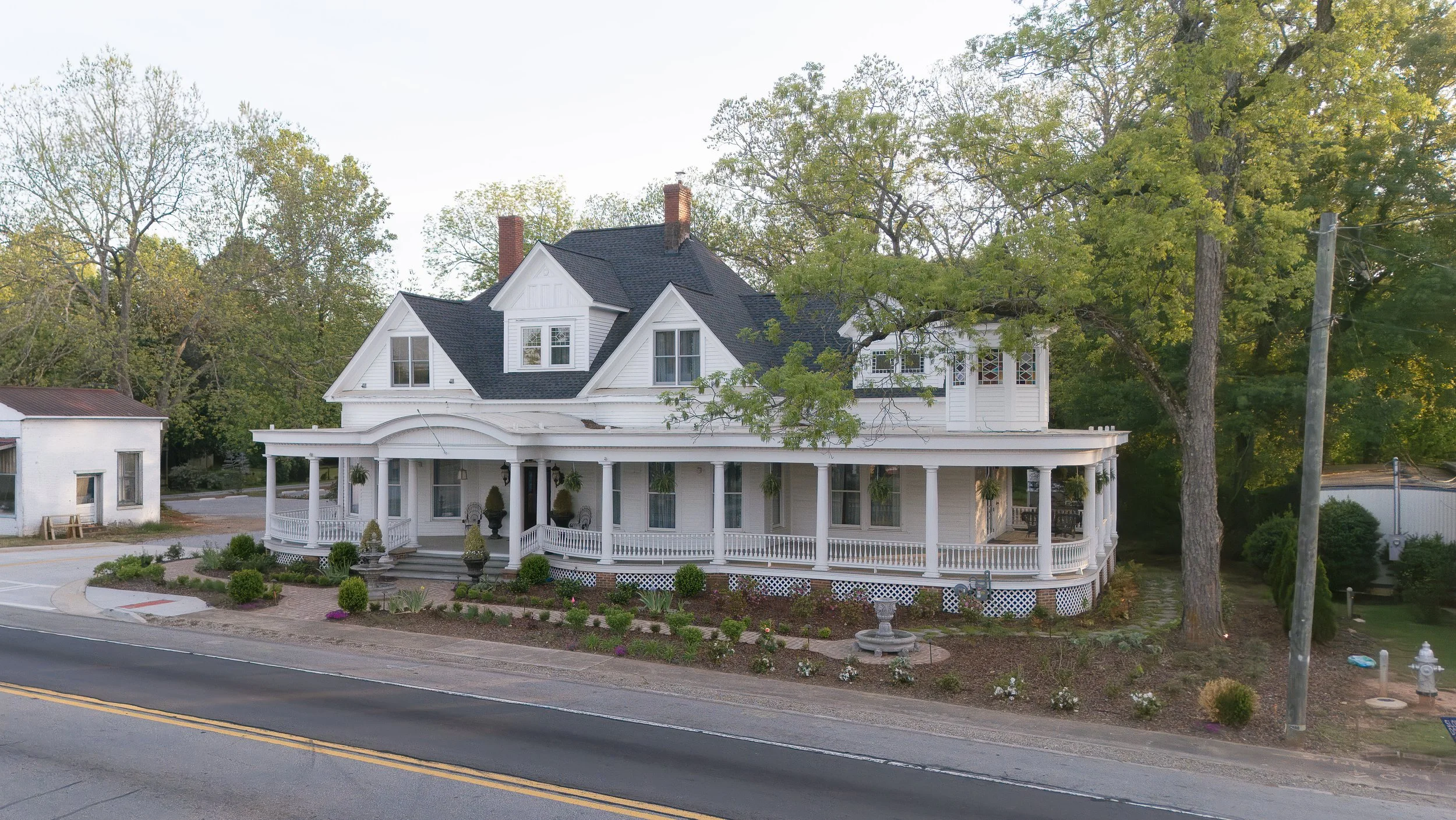 A large white Victorian house with a wraparound porch, dark roof, and multiple gables, surrounded by trees and a landscaped garden.
