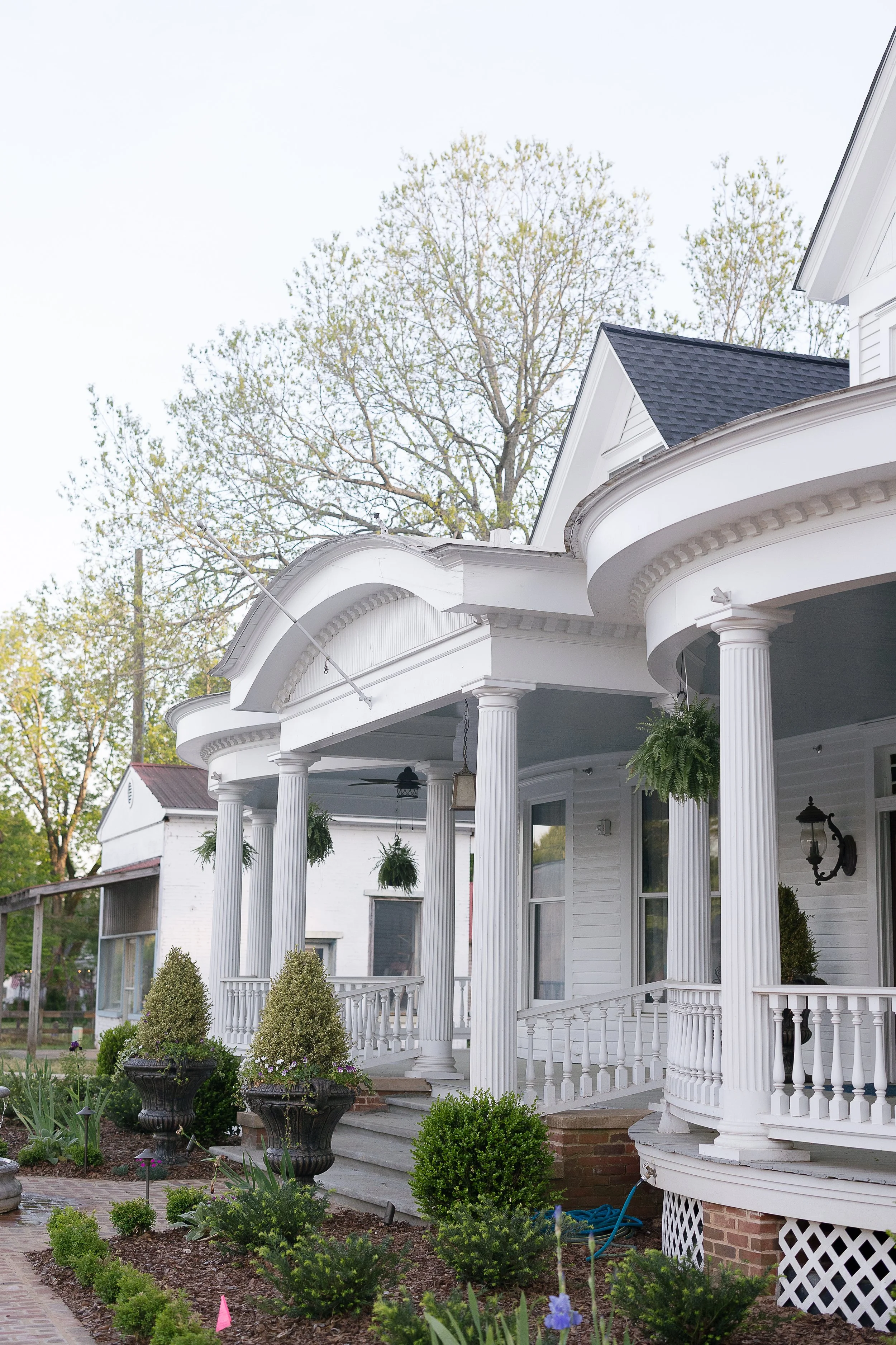 A white, Victorian-style house with a curved porch, white columns, and hanging plants. The house is surrounded by a garden with bushes, flowers, and potted plants, and has trees in the background.