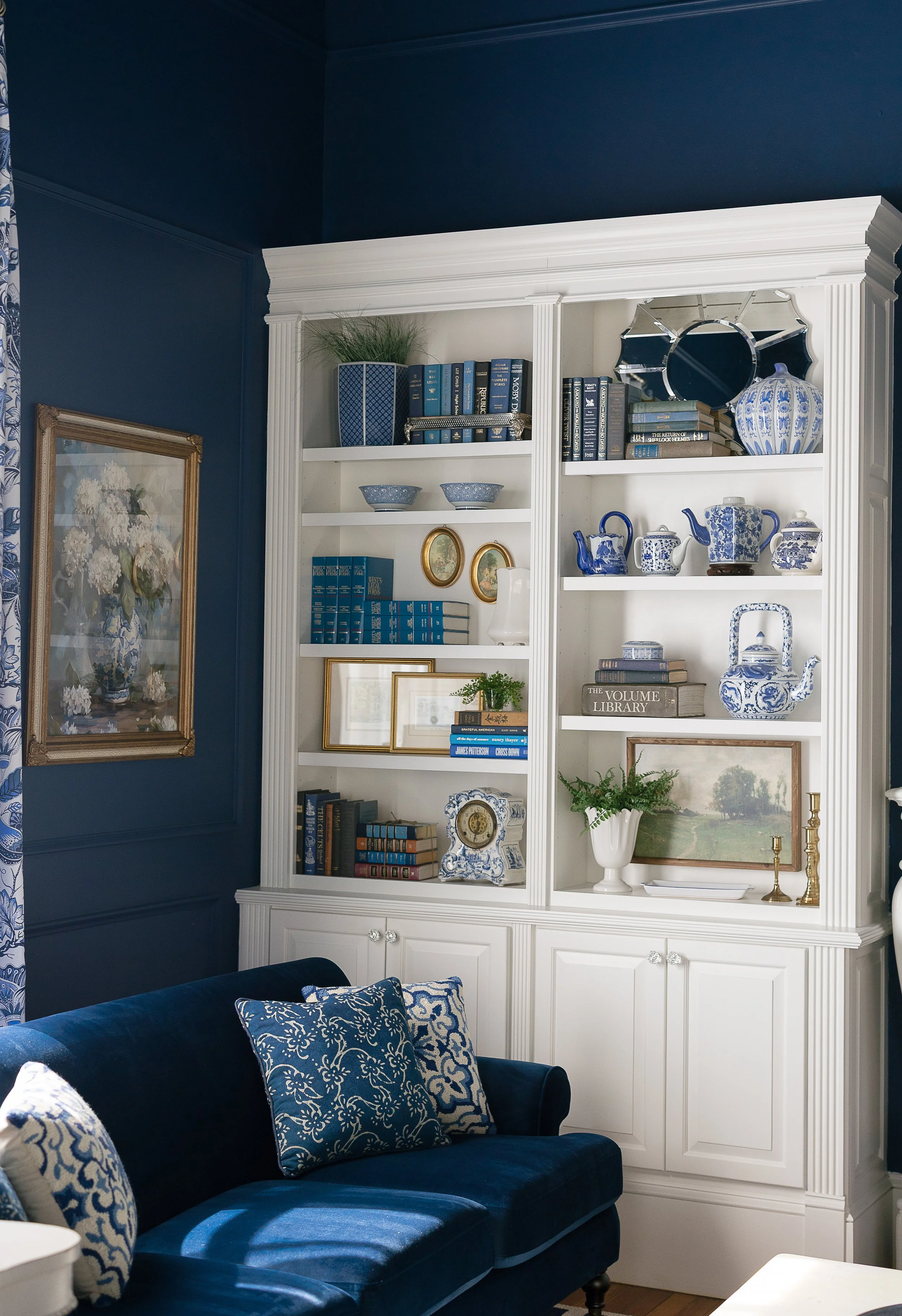 Living room corner with dark blue velvet sofa and decorative pillows, white built-in bookshelf with blue and white porcelain and books, dark blue walls, and framed art.