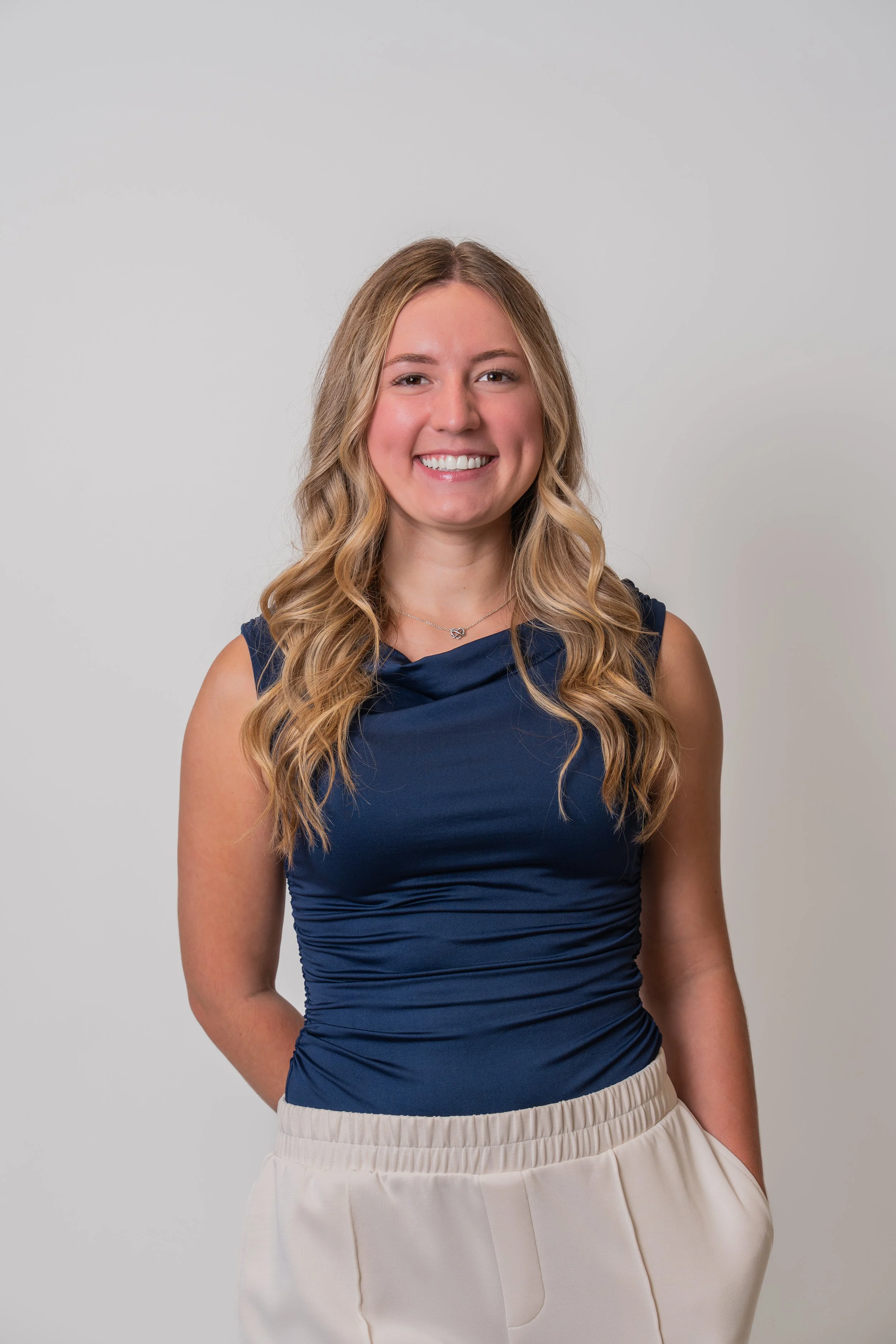 A young woman with long blonde hair smiling, wearing a navy sleeveless top and beige pants, standing against a plain light gray background.