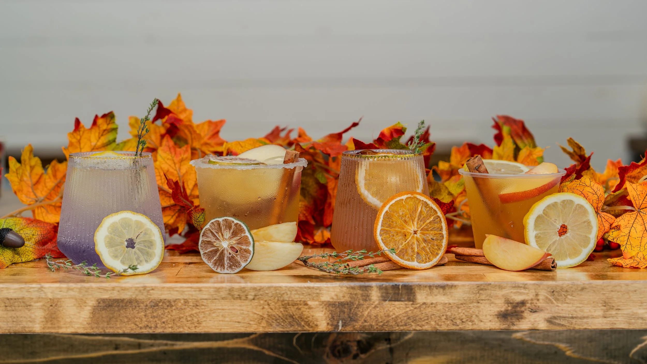 Four glasses of iced drinks with lemon slices and garnishes on a wooden table, surrounded by colorful autumn leaves.