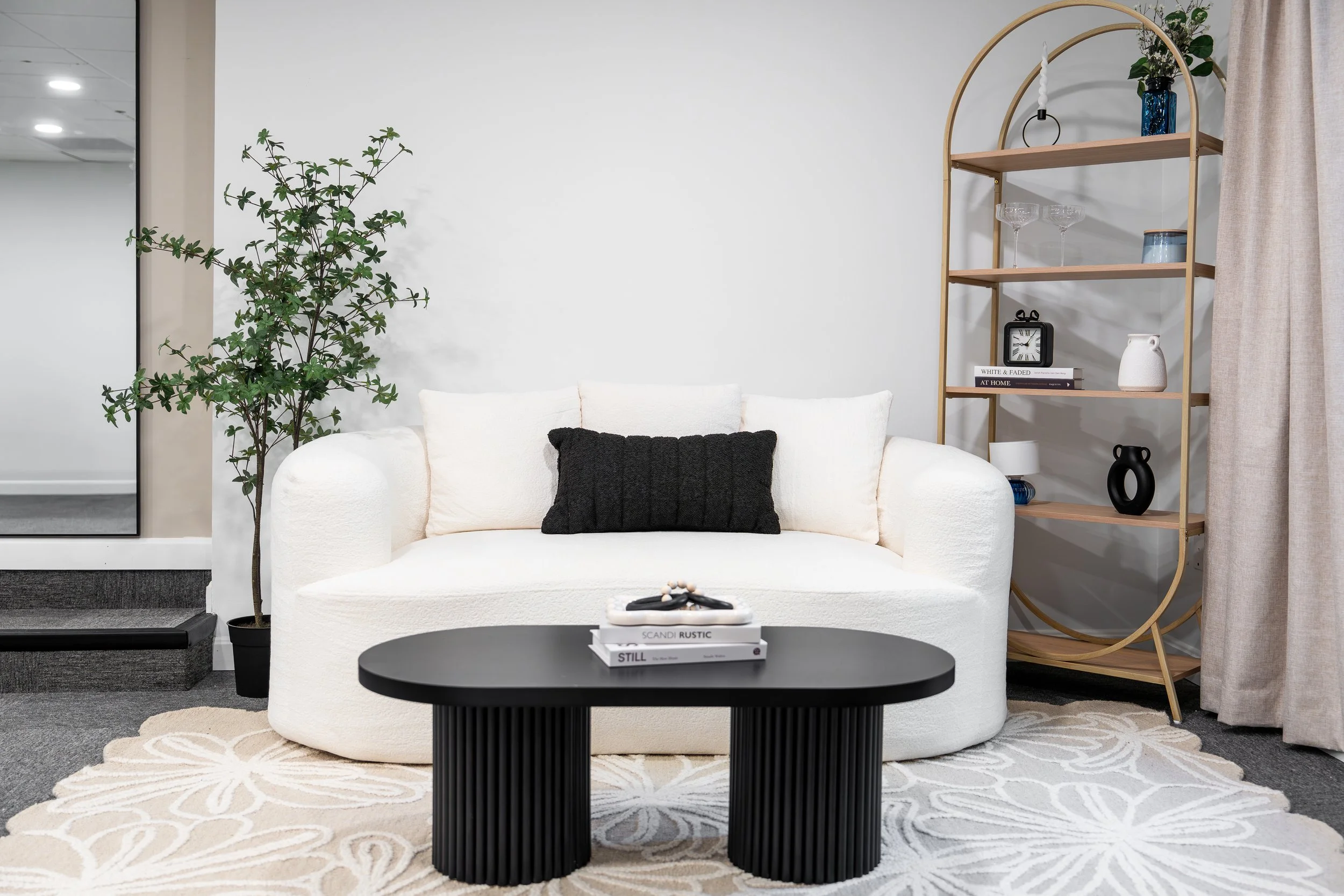 Modern living room with a white sofa, black pillow, black coffee table, tall green plant, gold bookshelf with decor, and a beige curtain.